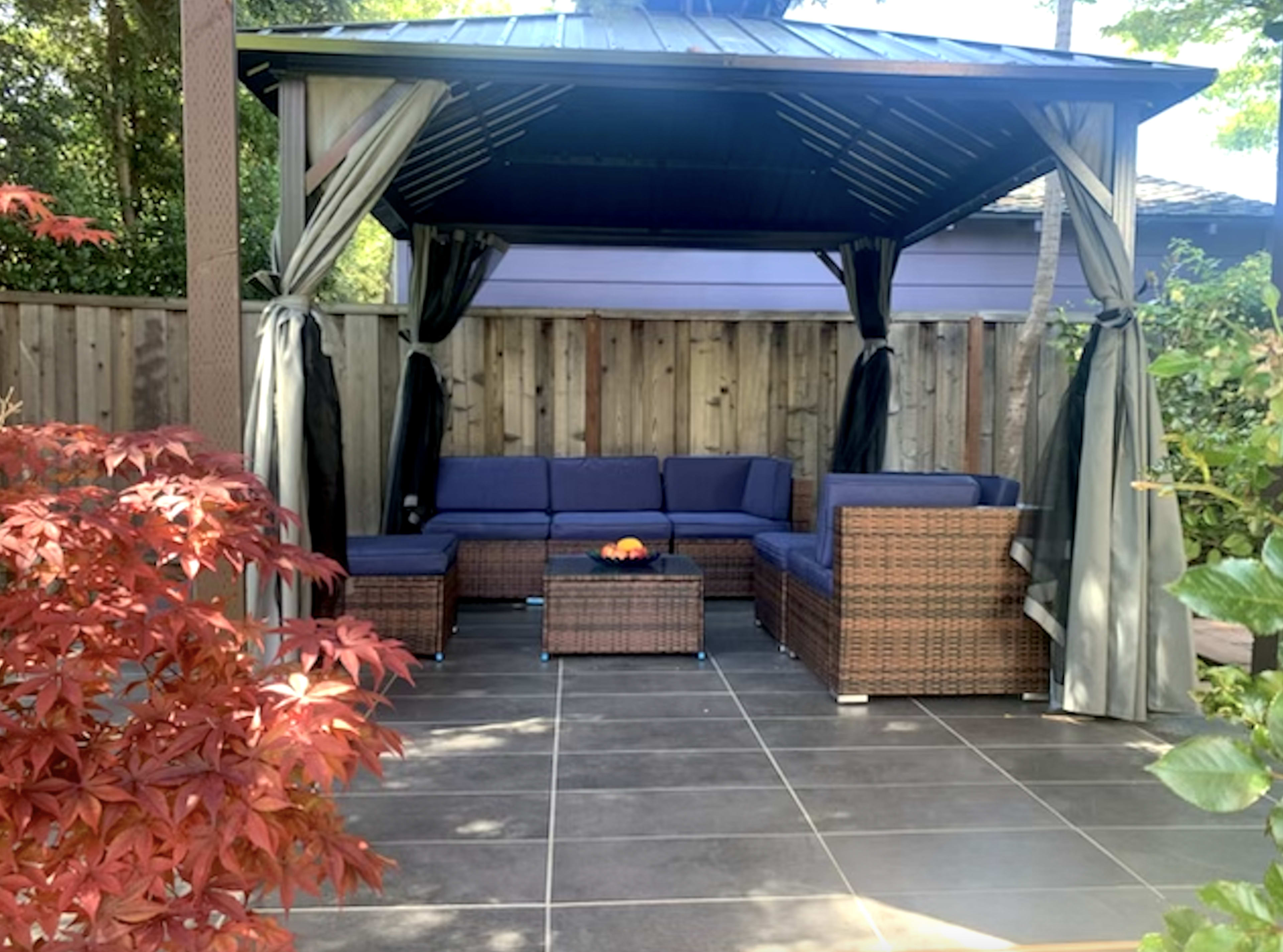A gazebo with a sectional seating area and a small table, surrounded by a wooden fence and colorful foliage.