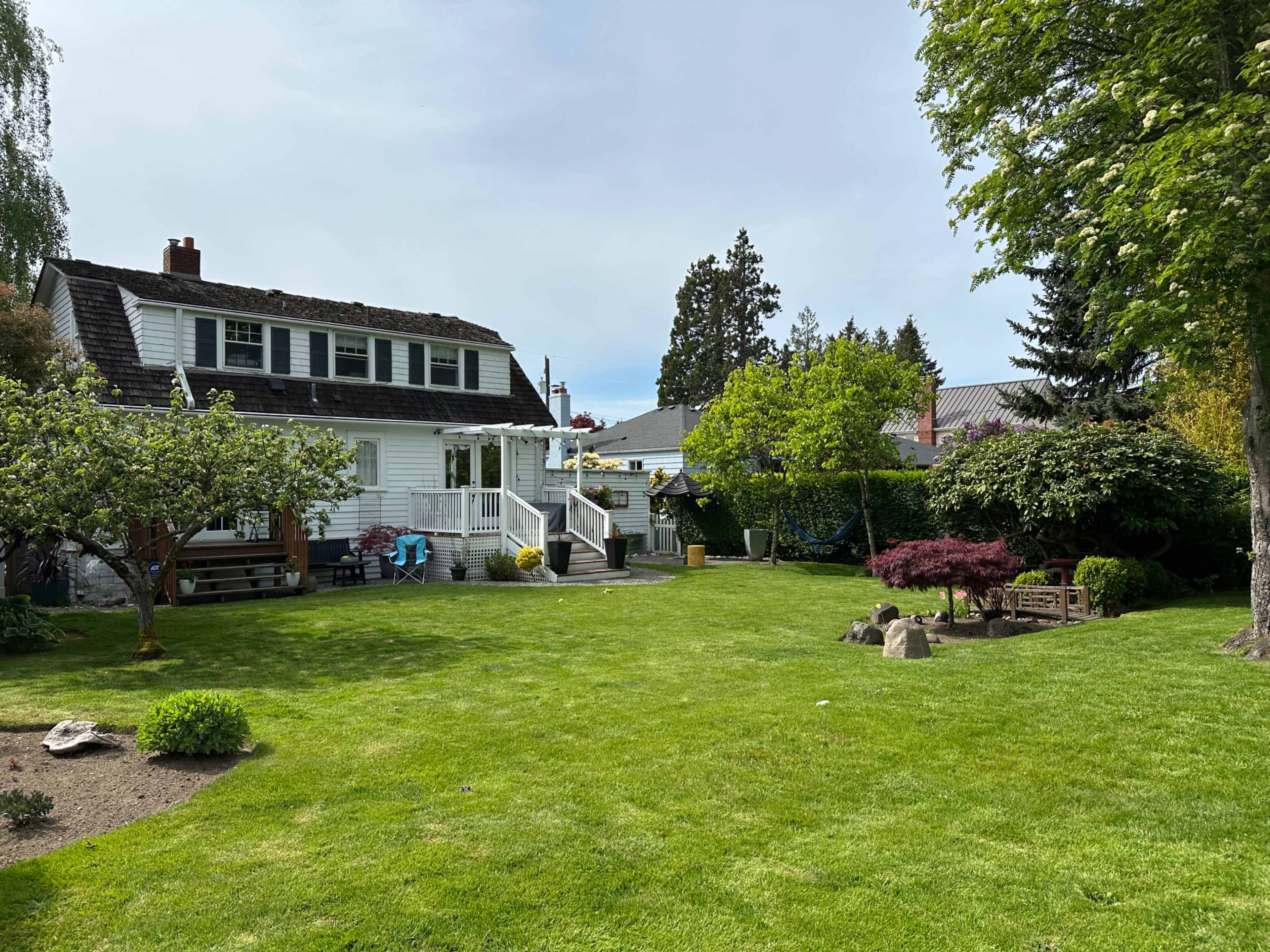The image shows a well-maintained lawn with a white two-story house in the background and a variety of trees and shrubs surrounding a garden area.