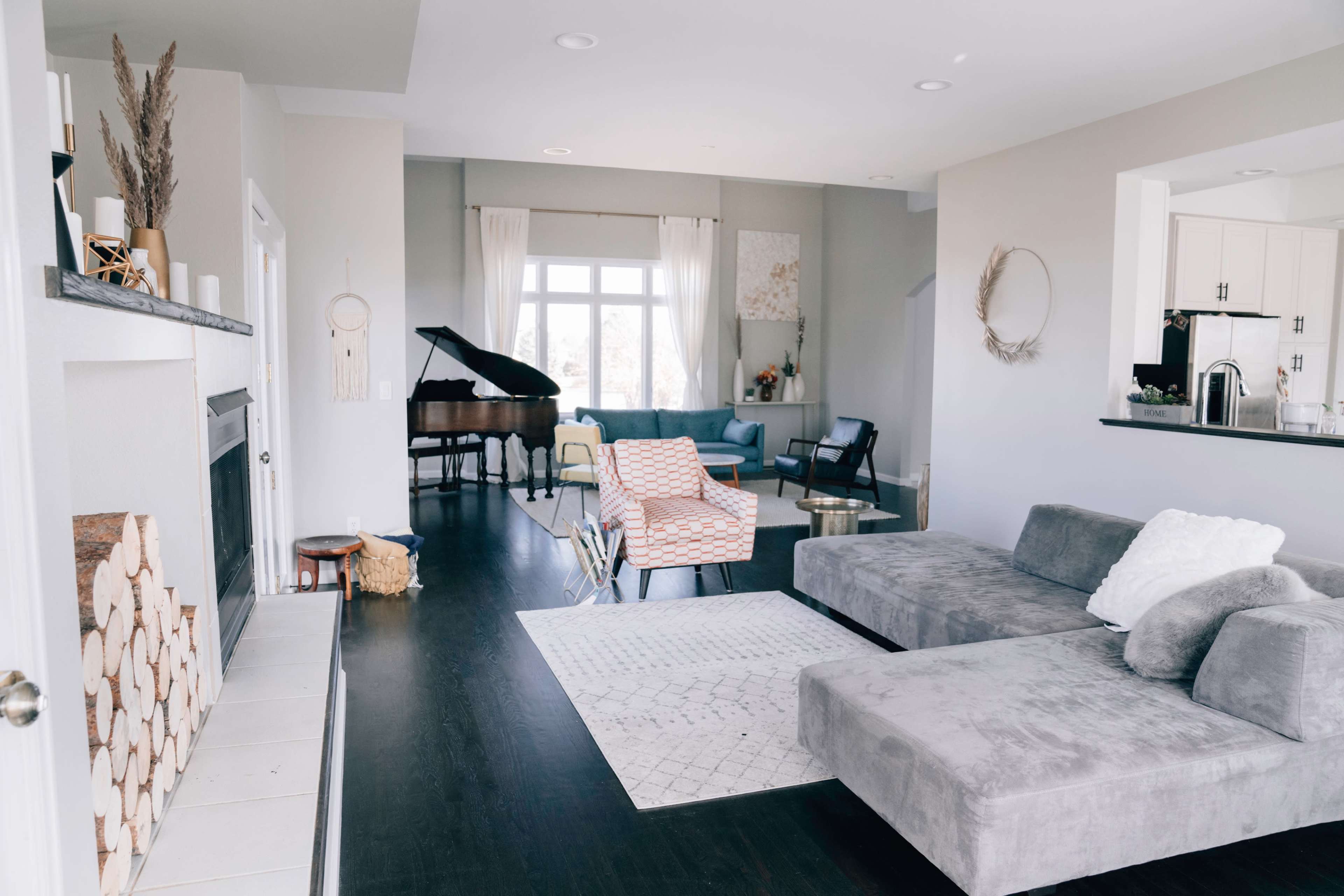 The image shows a modern living room with a sectional sofa, a piano, and a warm color scheme, featuring a wood-stacked fireplace and large windows.