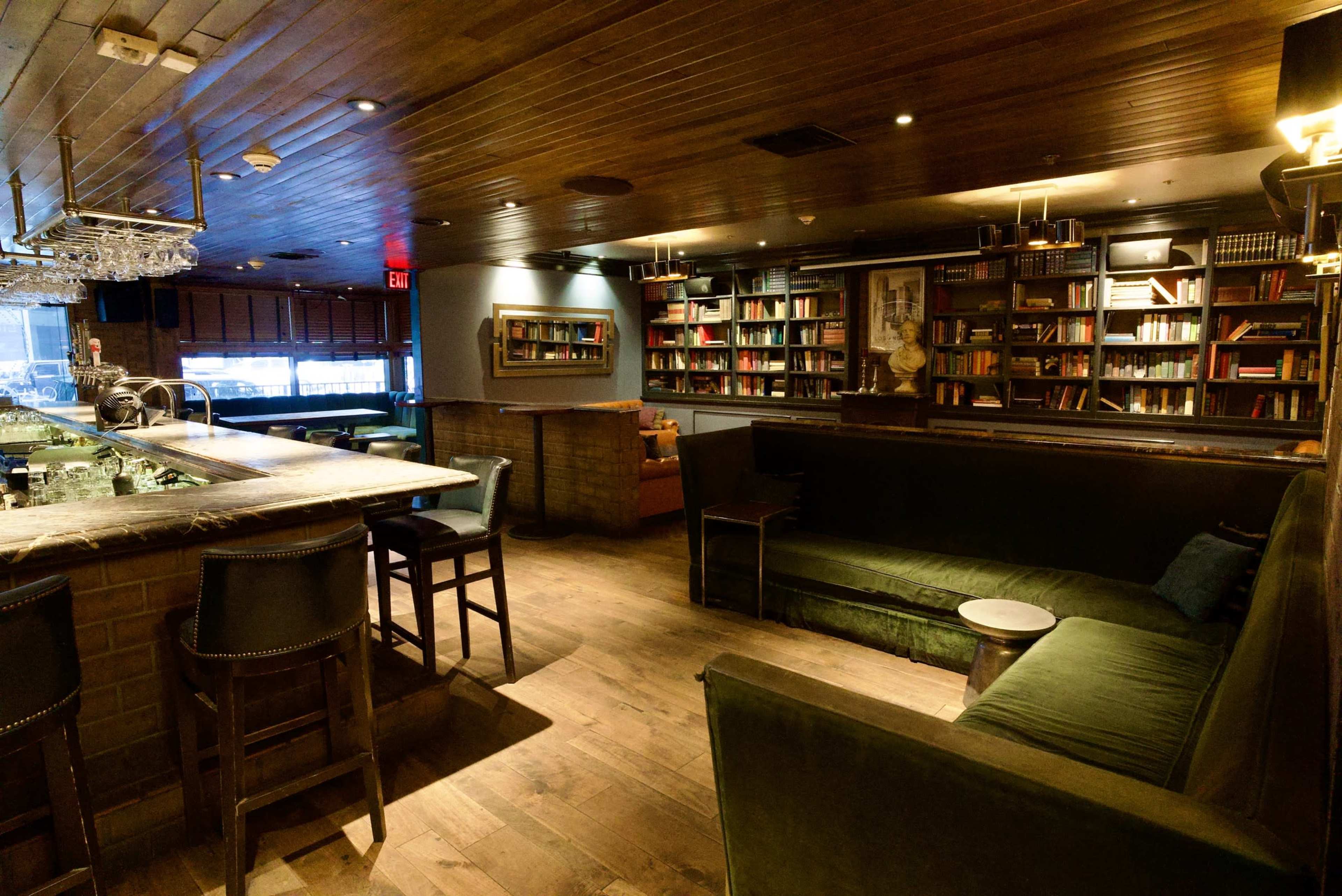 The image shows a dimly lit lounge area featuring a bar with high stools, a bookshelf with numerous books, and a green sofa against the wall.