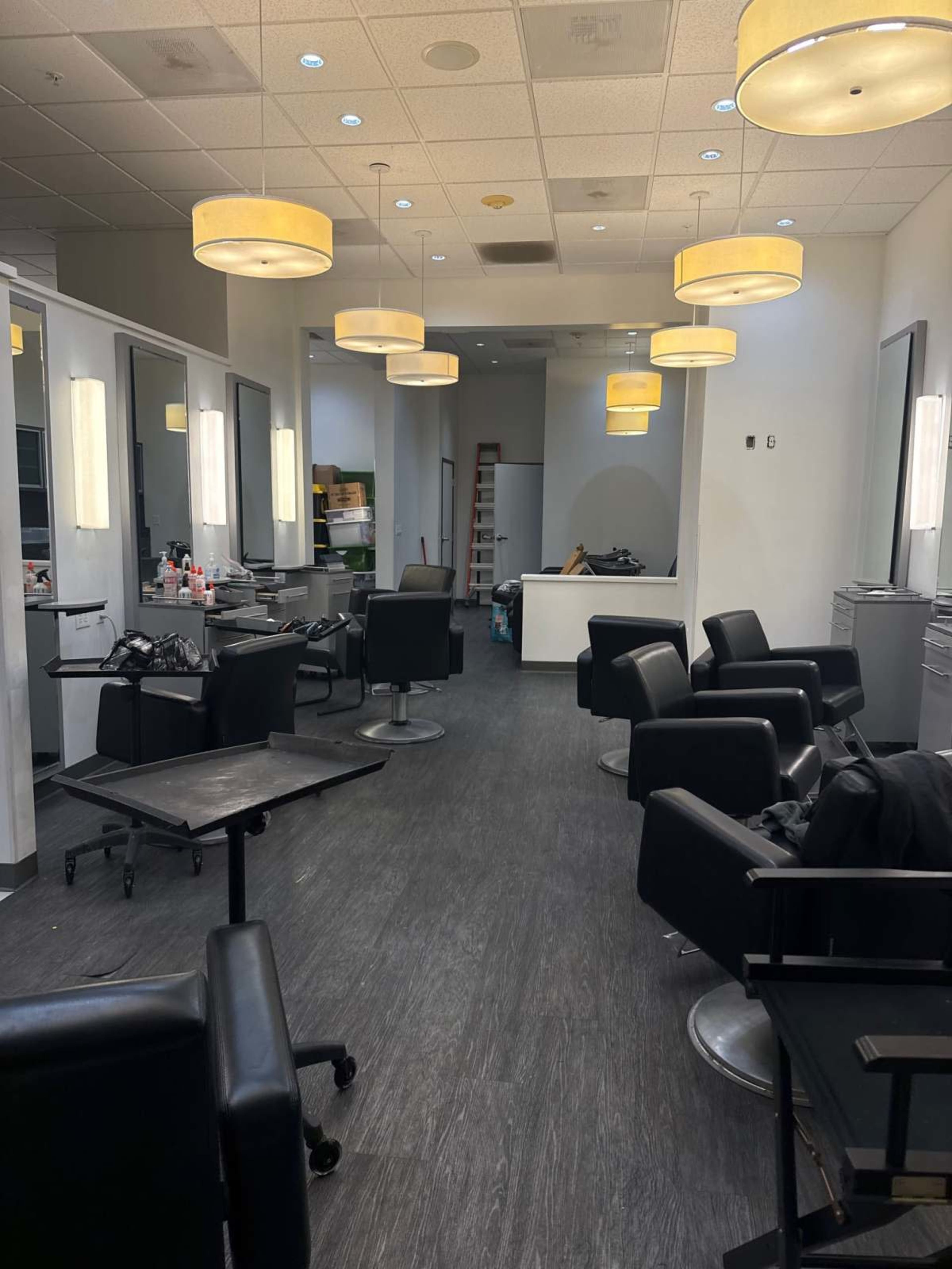 The image shows the interior of a hair salon with several black salon chairs, illuminated by pendant lights, and a reception area in the background.