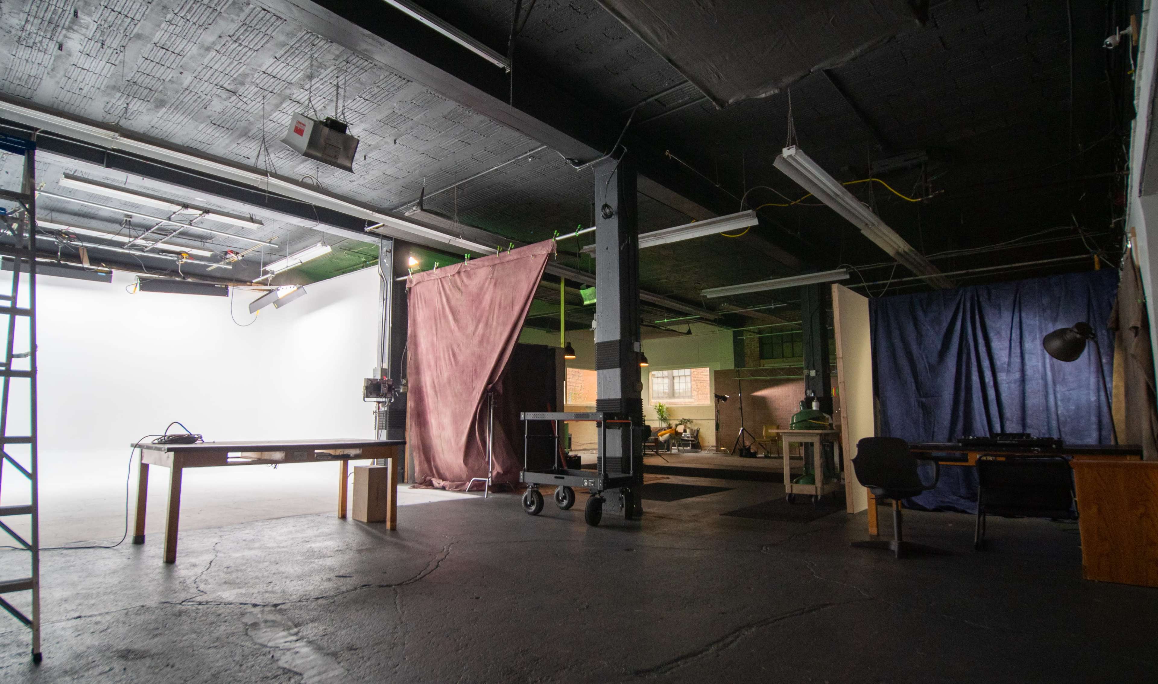 An industrial studio space with a white backdrop, tables, and curtains, illuminated by overhead lights.