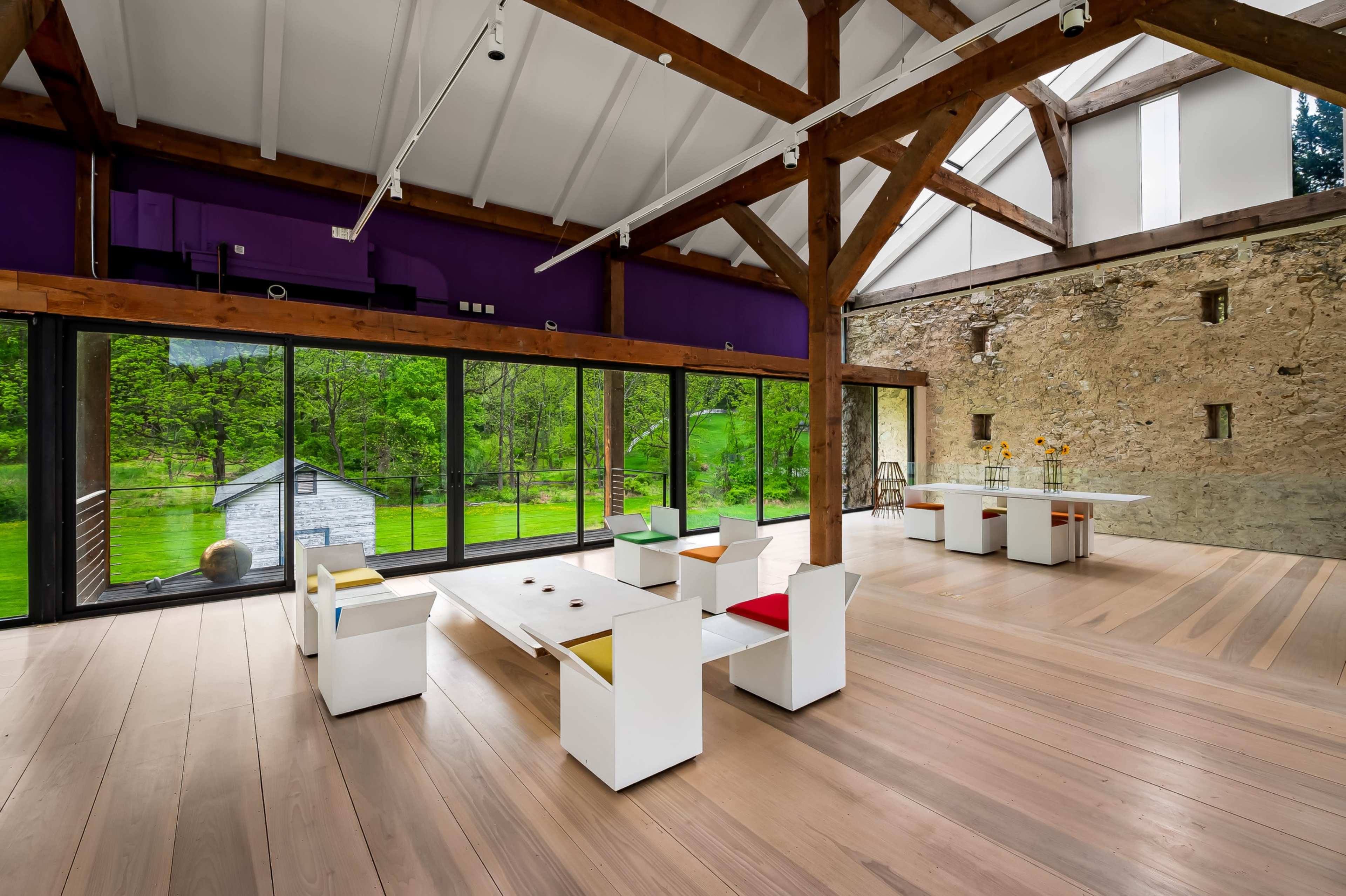 A spacious interior features a large open area with wooden beams, large windows overlooking greenery, and minimalistic white furniture.