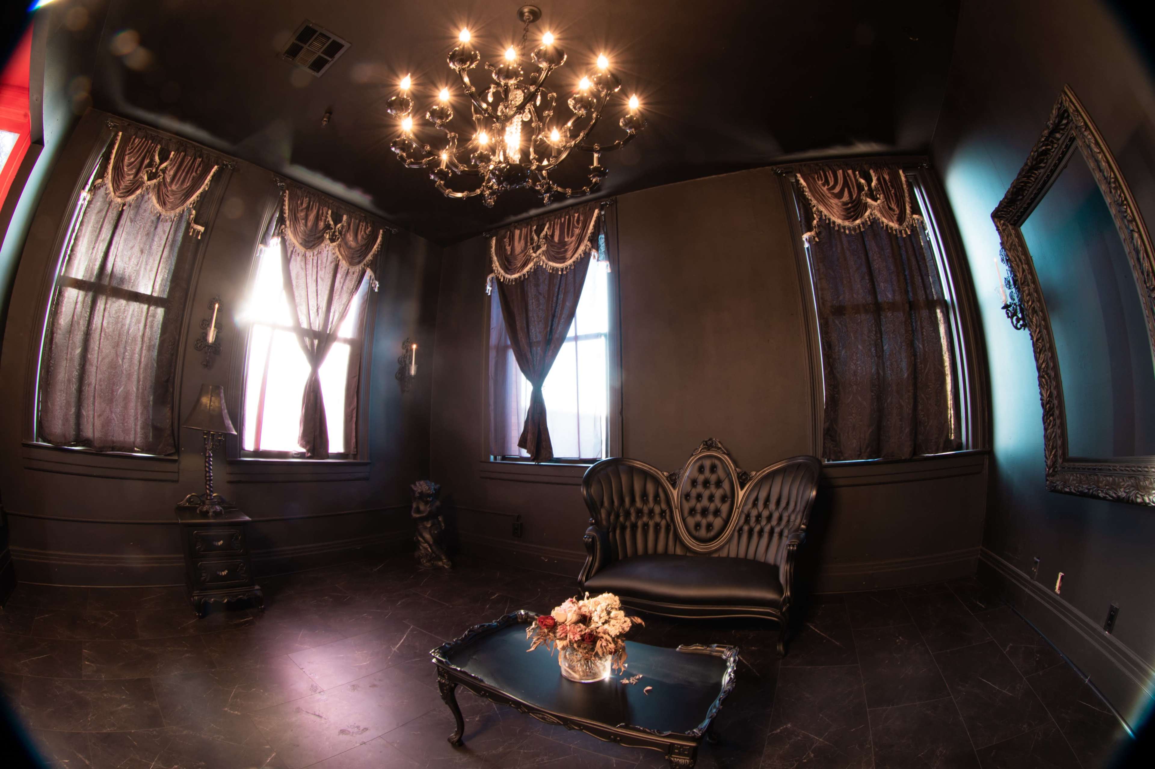 The image depicts a darkly decorated room featuring a vintage black sofa, an ornate chandelier, and large windows with heavy drapes.