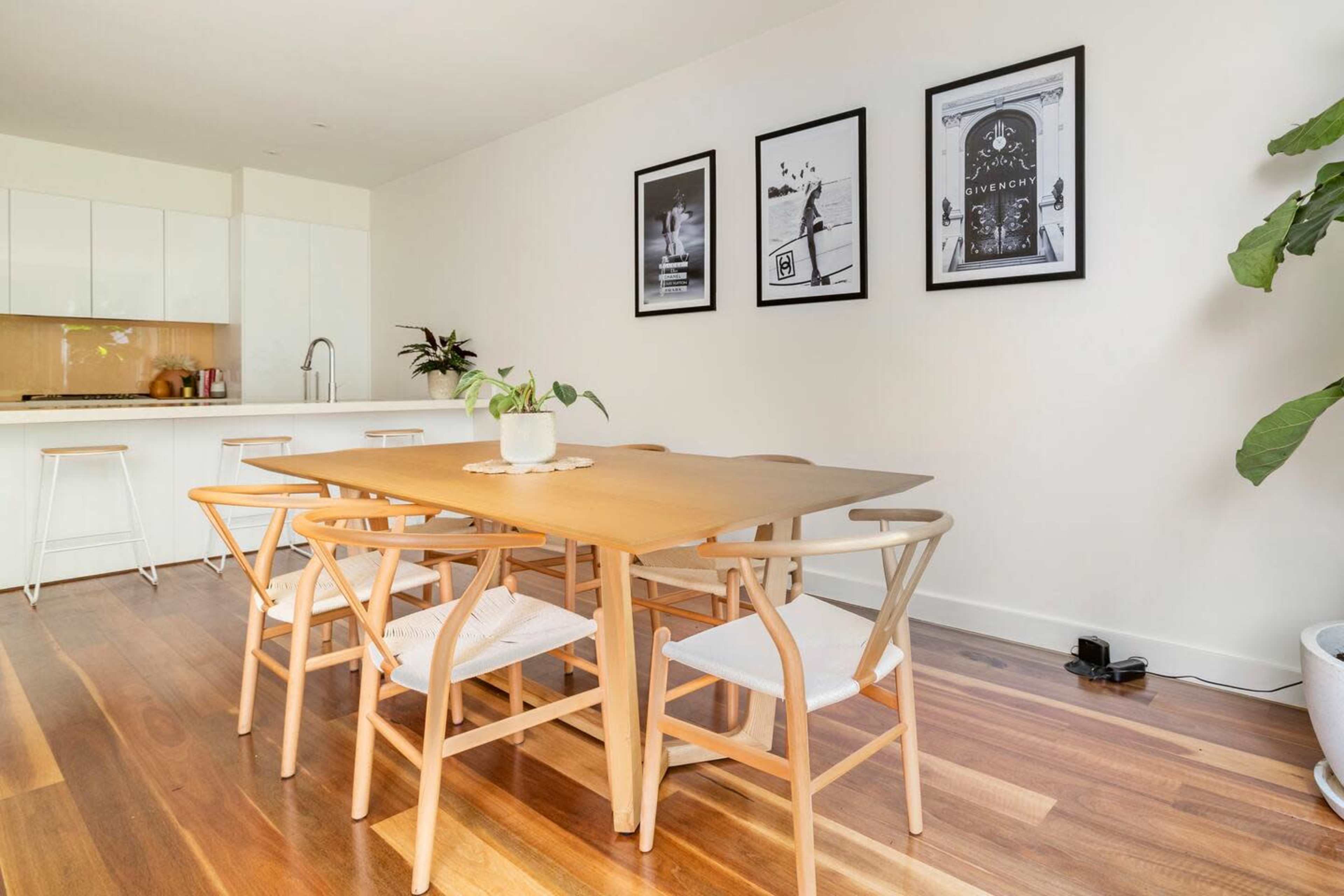 A modern dining area features a wooden table surrounded by six chairs, with three framed black and white photos on the wall and a plant in the corner.