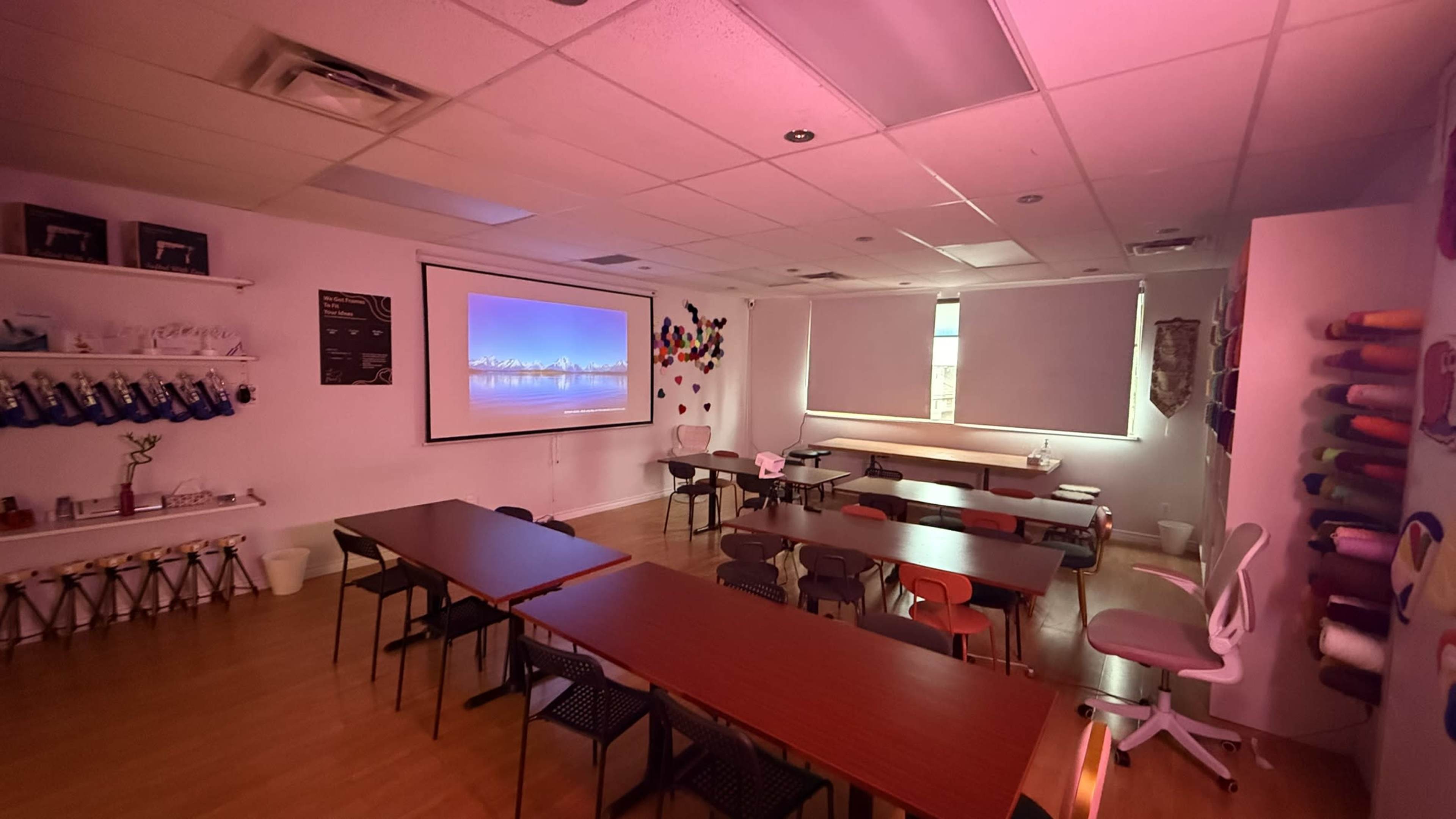The image shows a spacious room arranged with several tables and chairs, featuring a projector screen at one end and large windows with natural light.