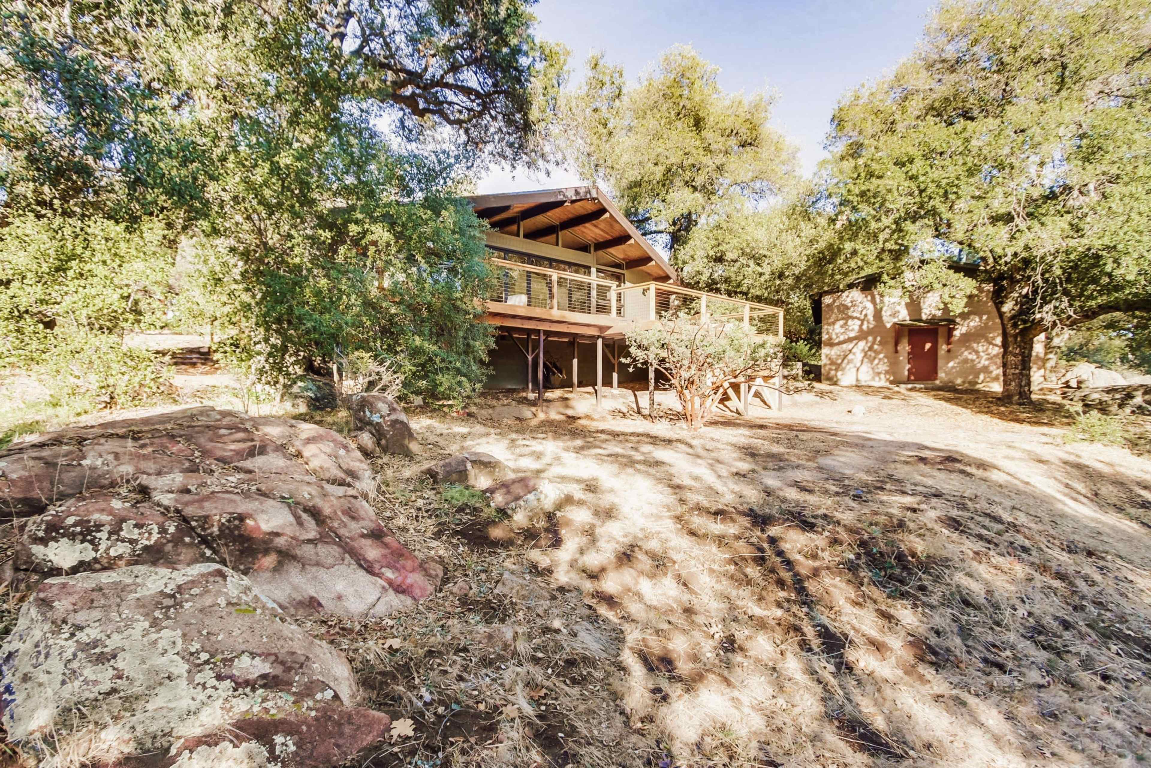 A wooden house sits on a sloped terrain surrounded by large trees and rocky outcrops.
