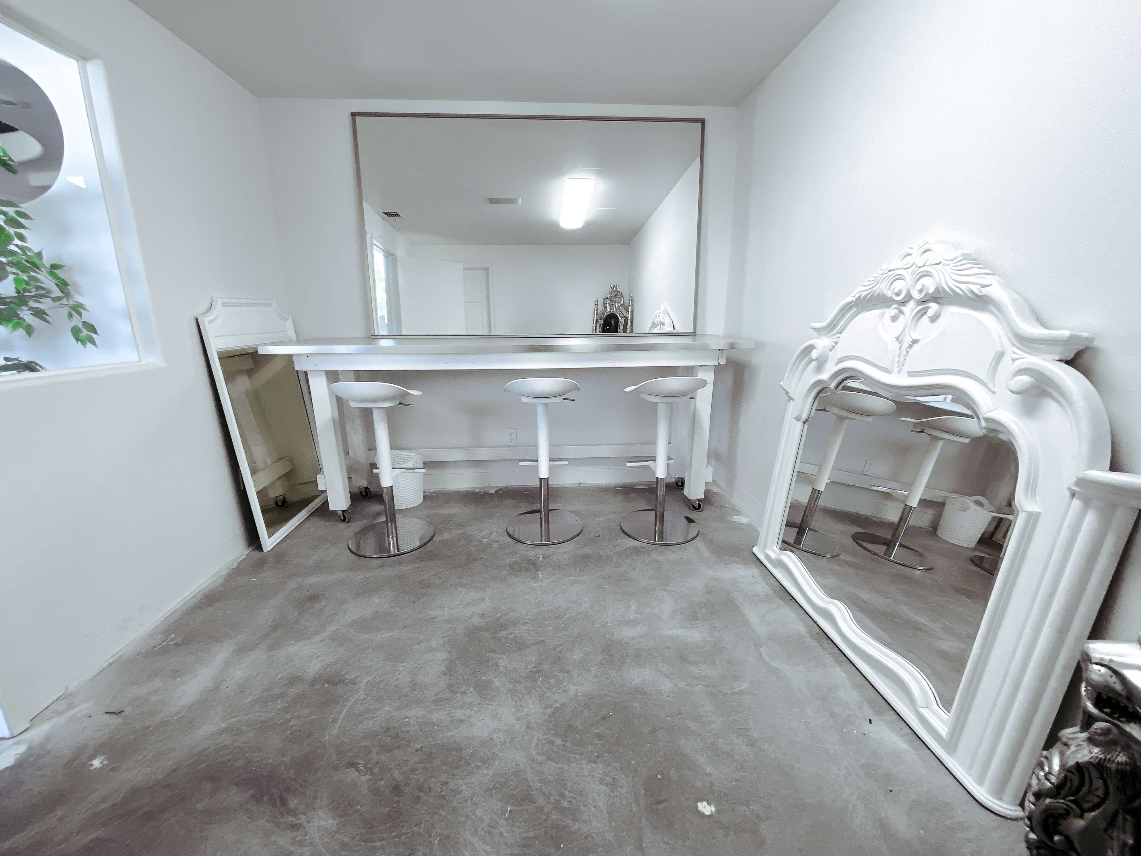 A minimalist room with a white bar counter and three white stools, accompanied by an ornate mirror and a decorative wall mirror.