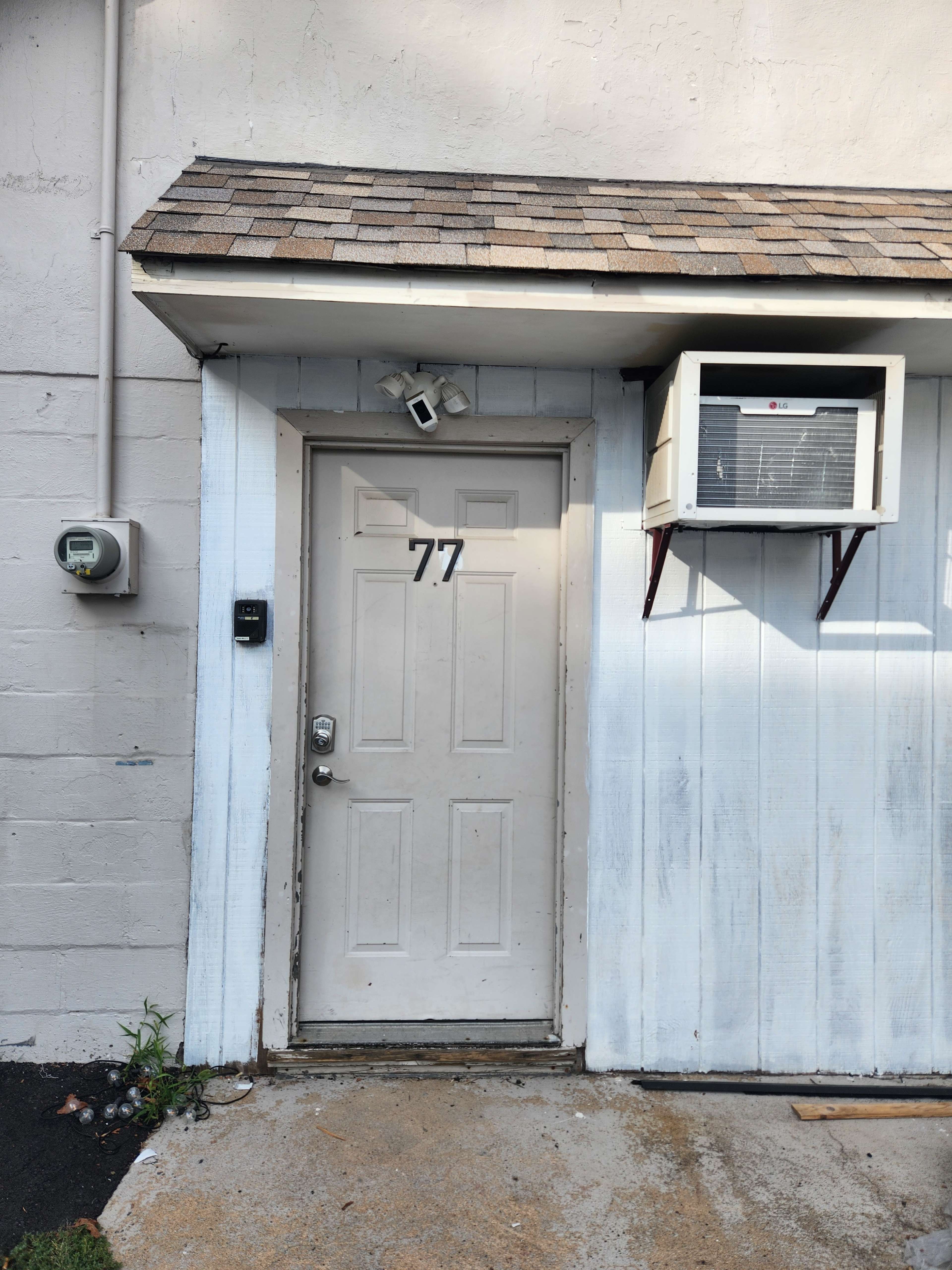 A white door with the number 77 on it, accompanied by a security camera, an air conditioning unit, and an electrical meter on the adjacent wall.
