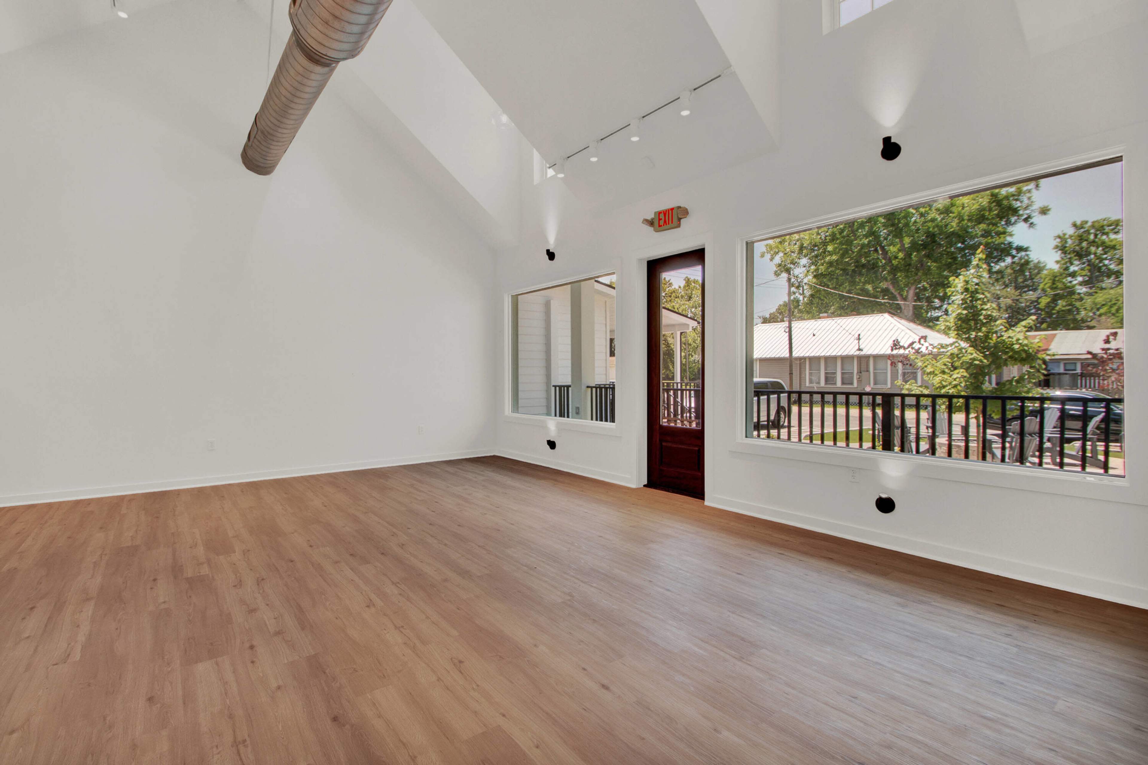 The image shows a bright, empty room with high ceilings, large windows, and light wooden flooring.