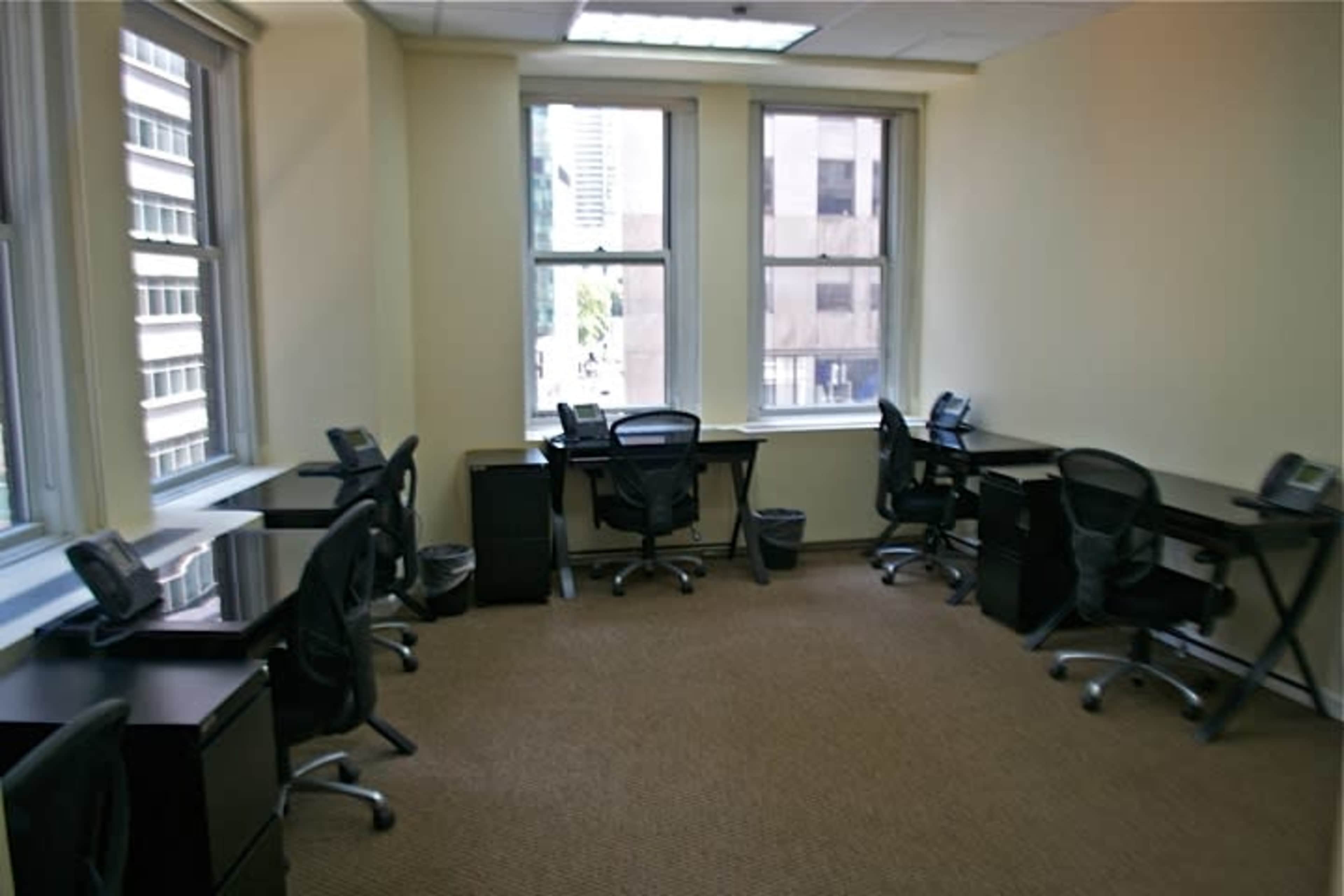 A vacant office space with five black desks and chairs arranged near large windows.