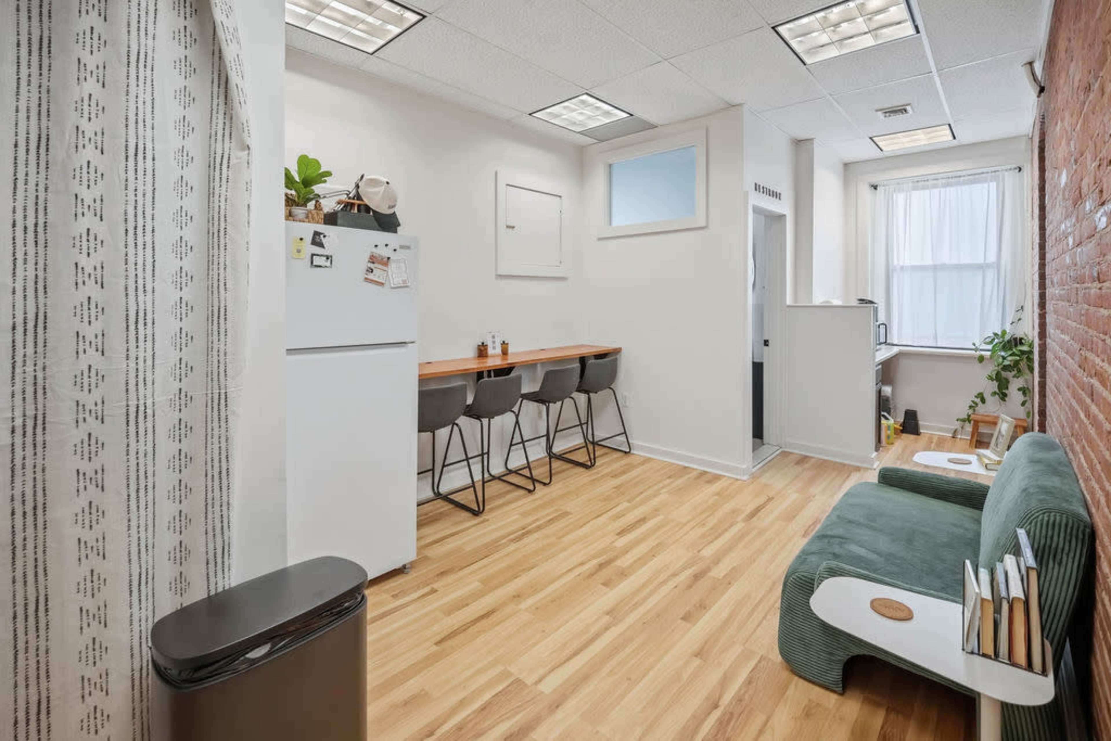 The image shows a modern, minimalist kitchen area with a small dining table, sleek chairs, and a refrigerator, all set against a backdrop of wood flooring and exposed brick walls.