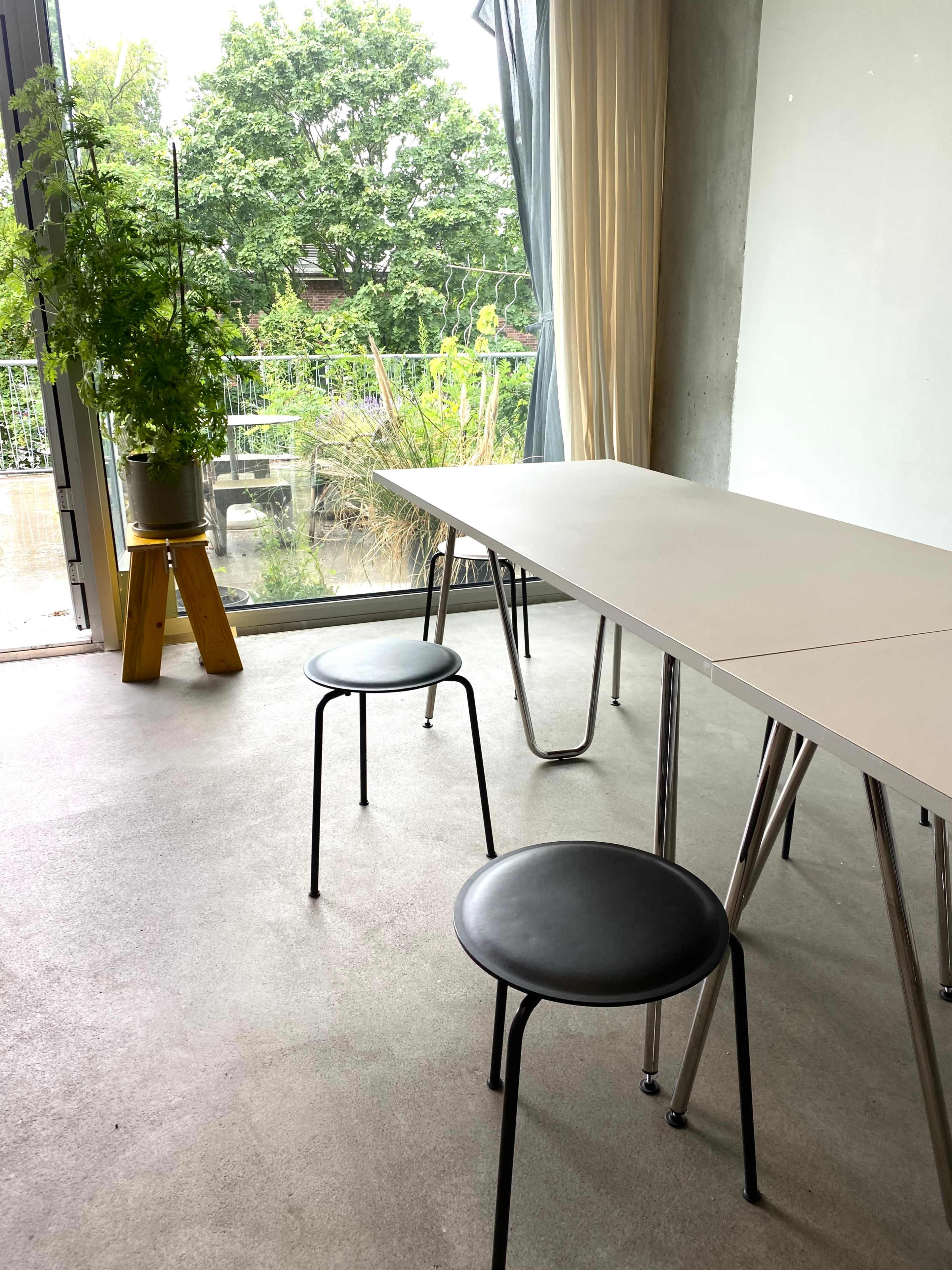 A modern interior features a simple table with two black chairs beside a large window overlooking a garden with greenery.