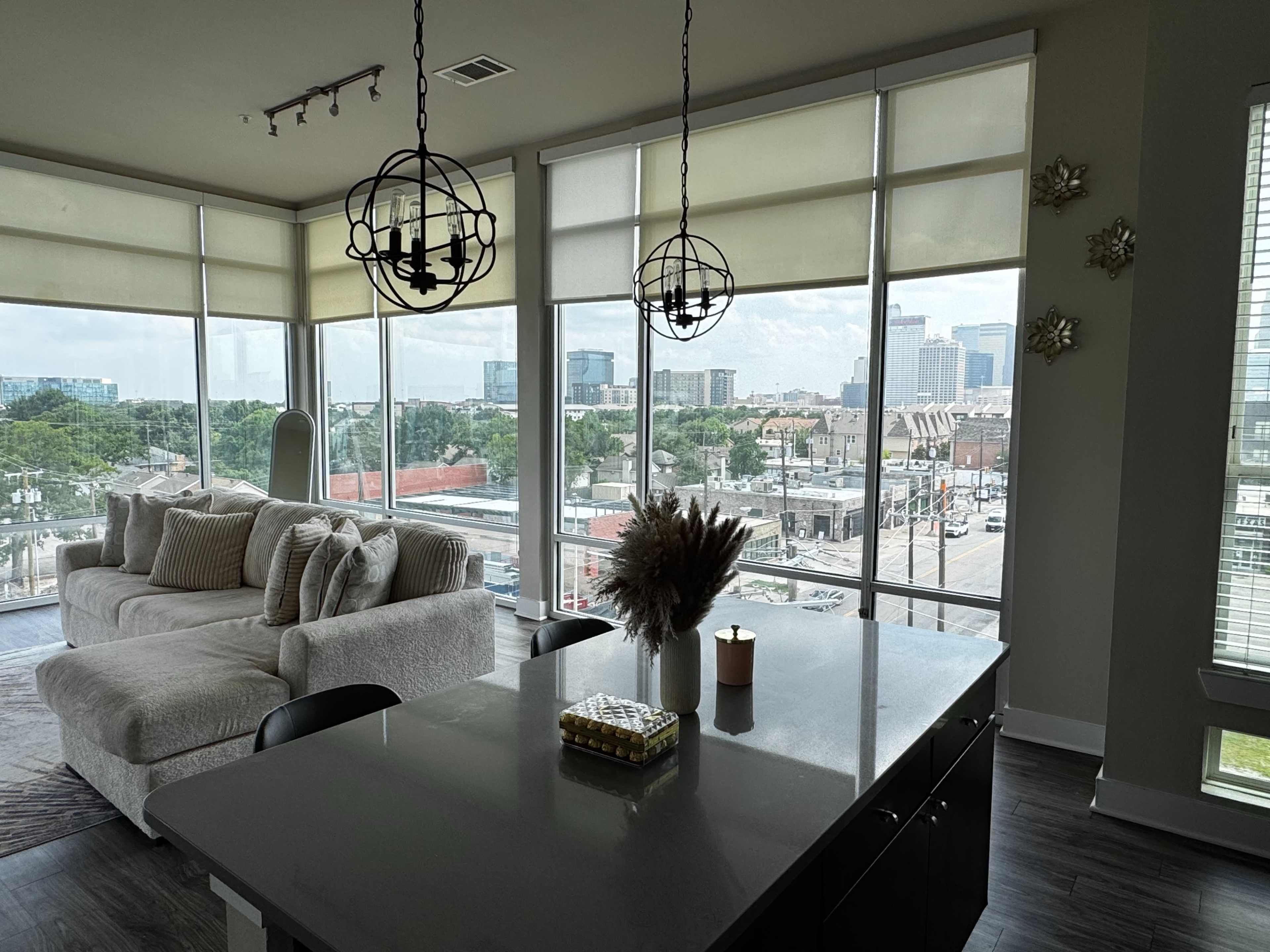 A modern living space features a light-colored sofa, a kitchen island, and large windows showcasing a view of a city skyline.