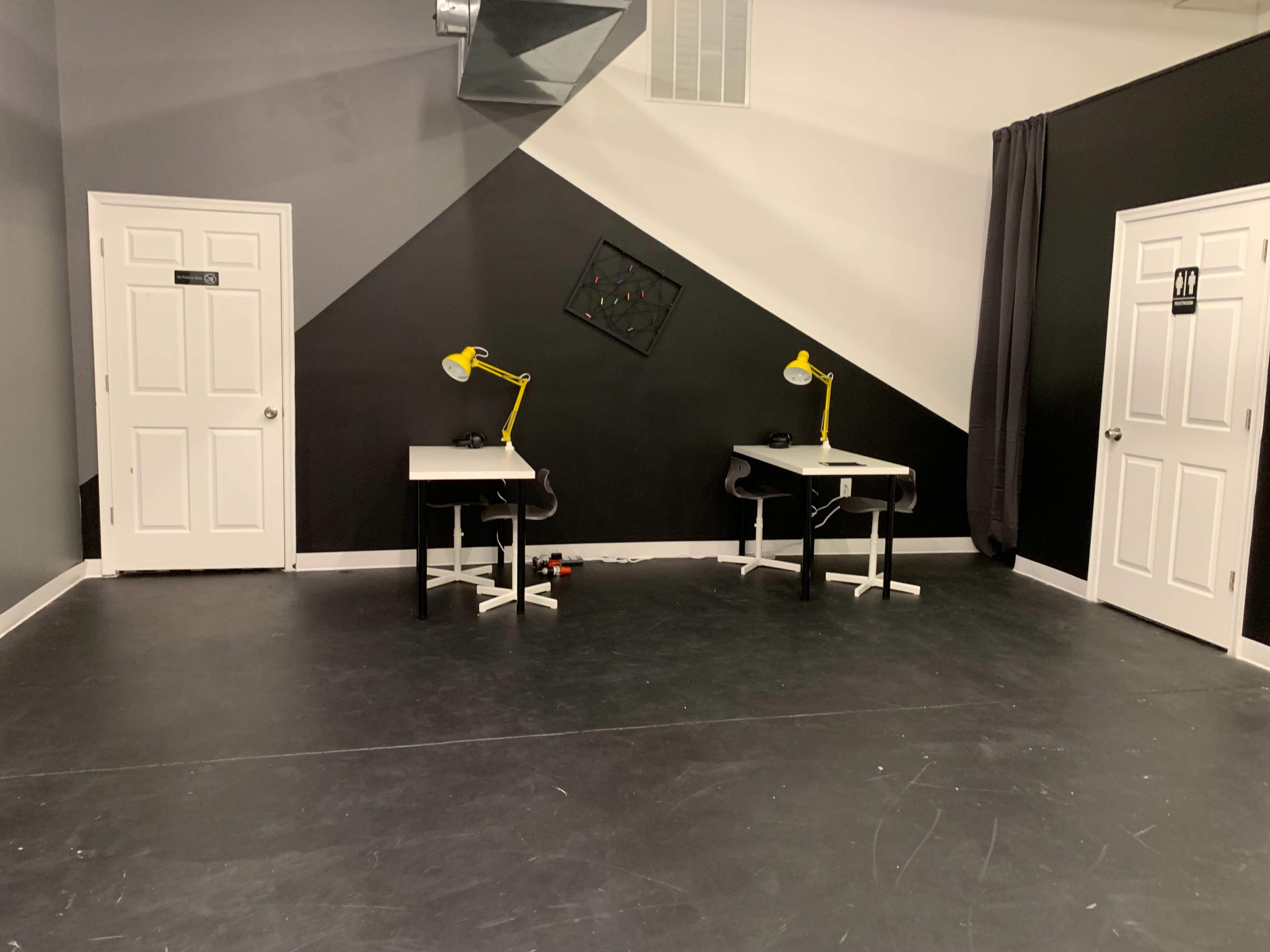 A minimalist workspace with two desks, each equipped with a yellow desk lamp, situated against a diagonal black and gray wall.