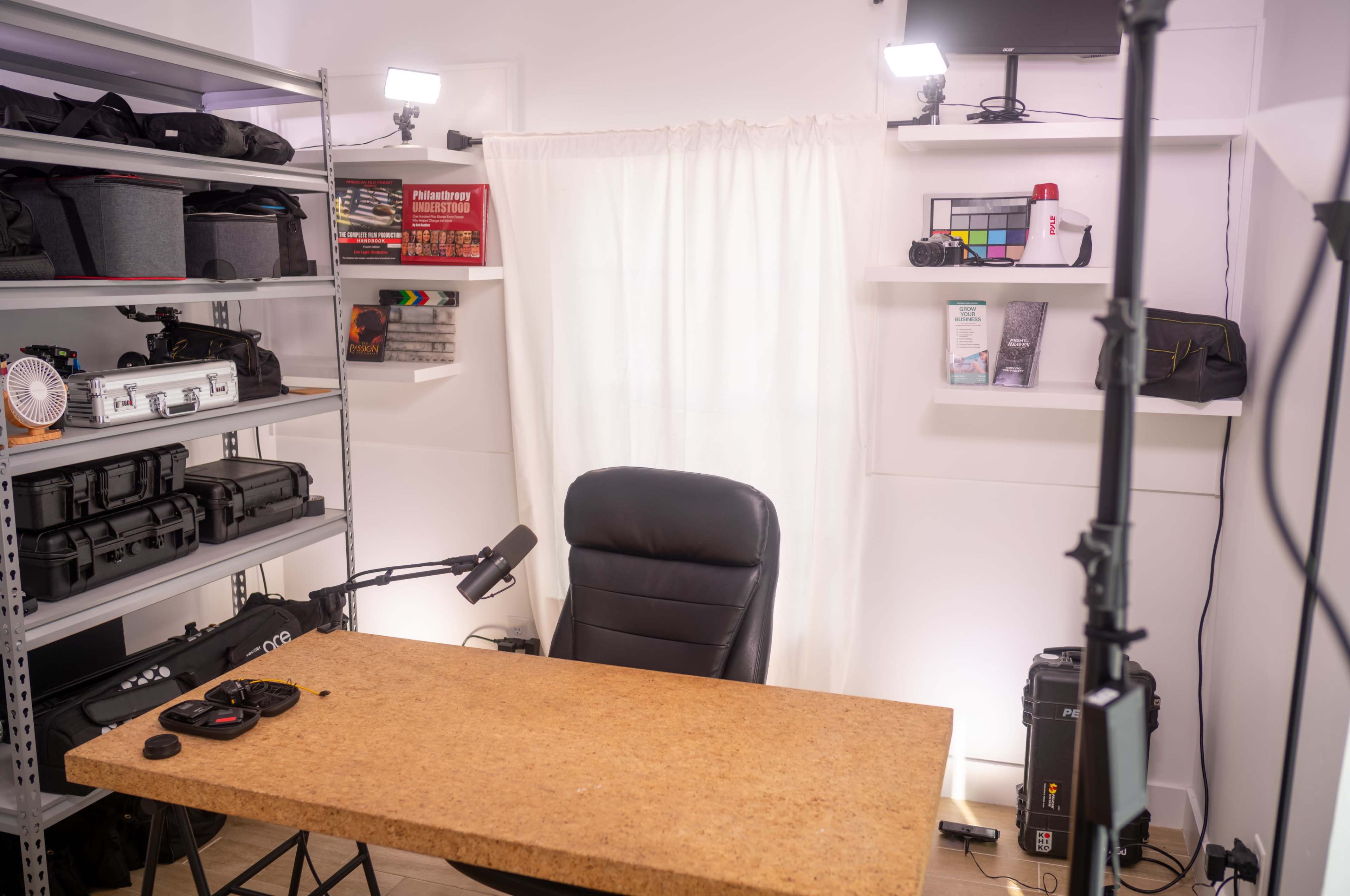 The image shows a well-organized workspace featuring a large cork table, a black chair, and shelves stocked with various equipment and supplies, illuminated by bright lights.