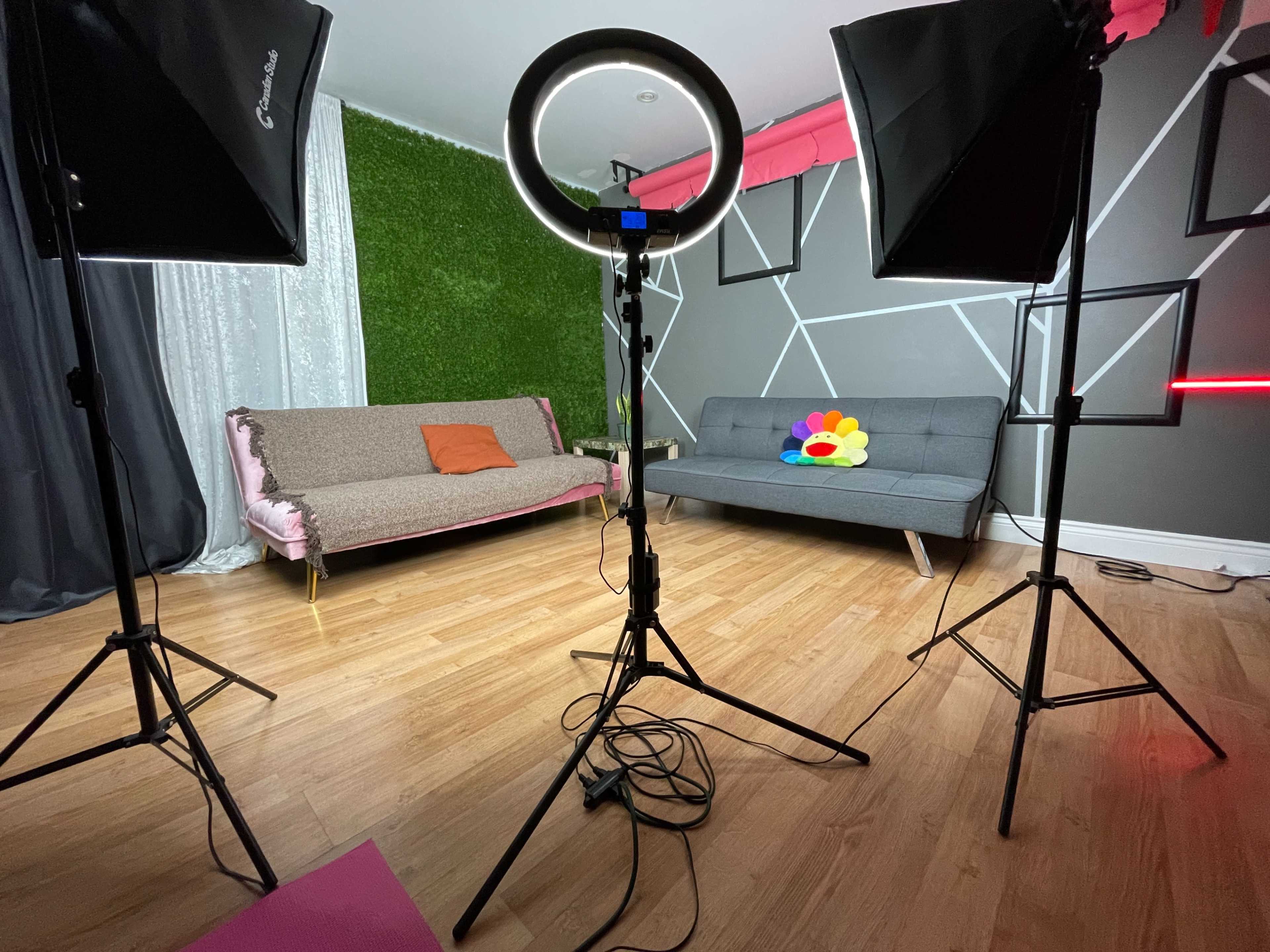 A modern studio setup featuring two softbox lights, a circular ring light, and two sofas arranged against a decorative wall.