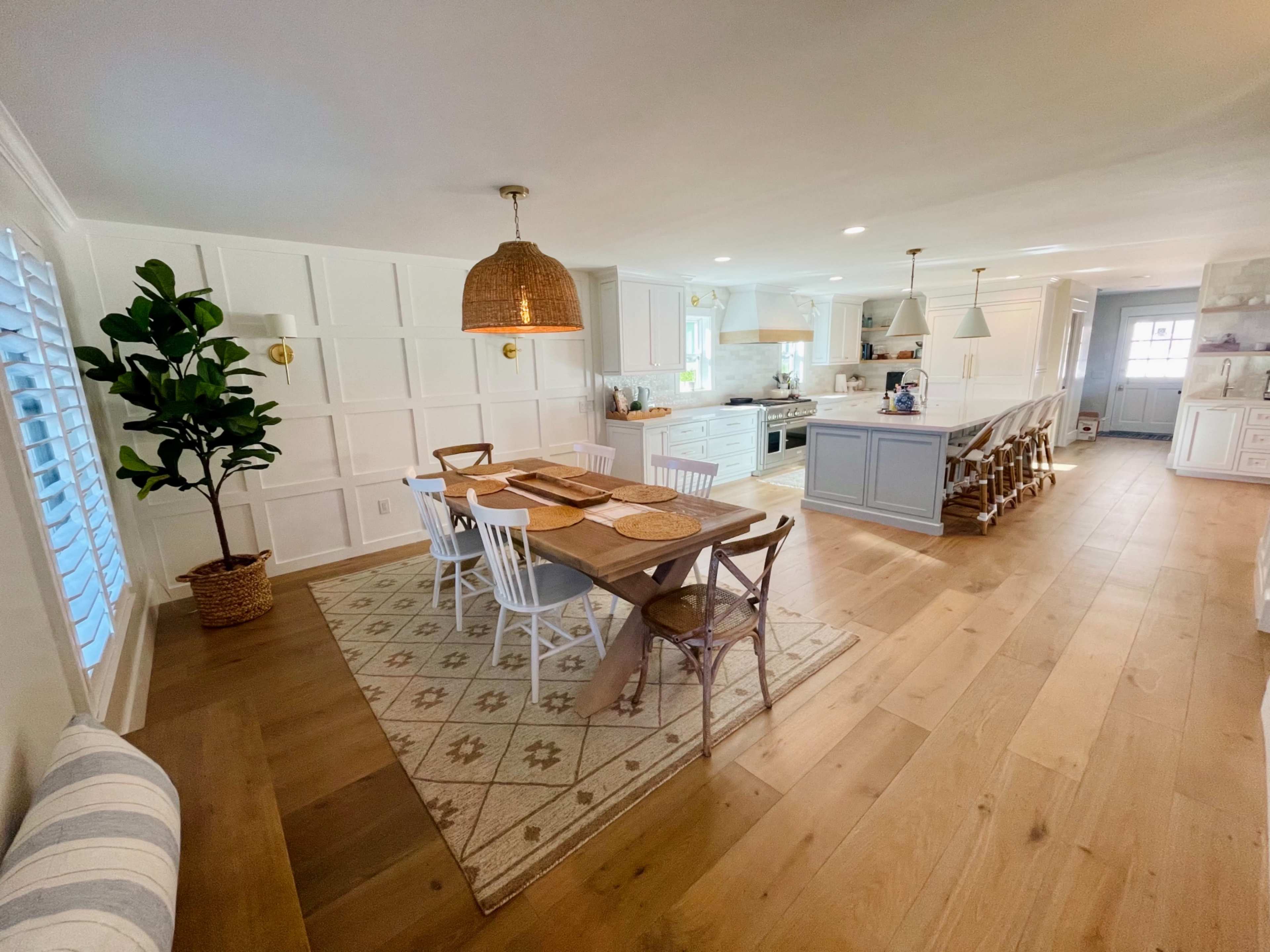 A bright, open kitchen and dining area features a wooden table with woven placemats surrounded by white chairs, a large island, and light-colored cabinetry.