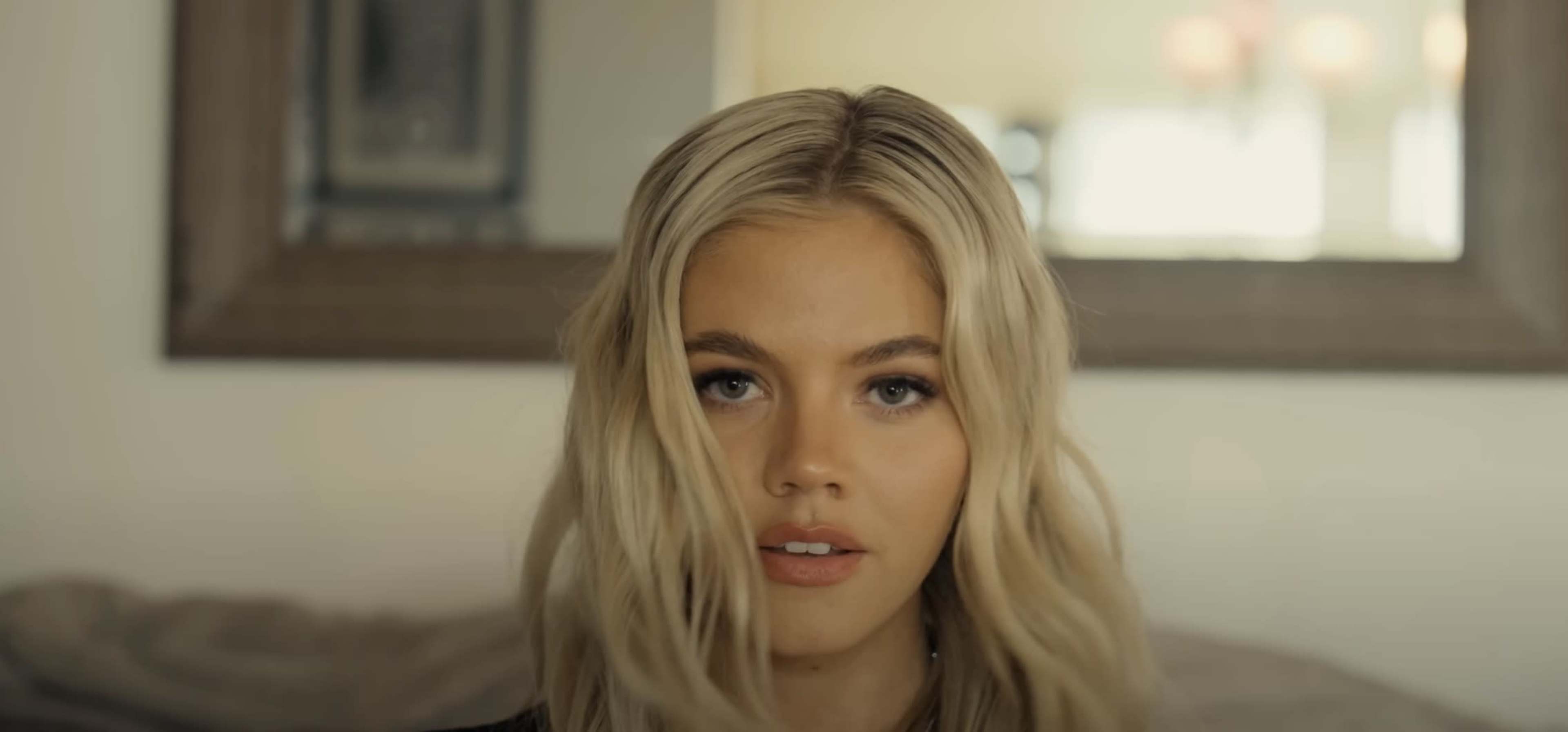 A person with long, wavy blonde hair is looking directly into the camera, set against a blurred background with a mirror and soft lighting.