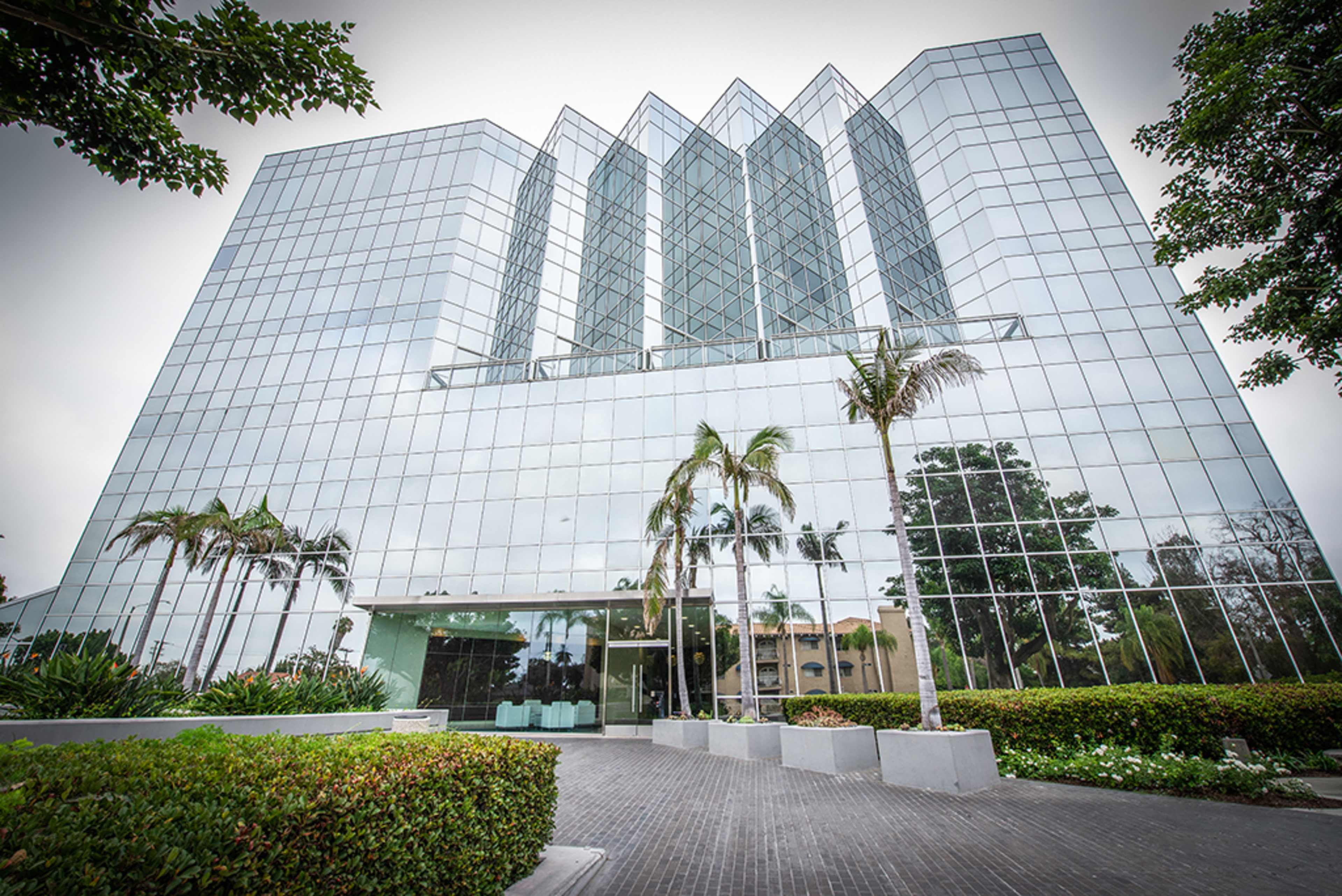 A modern glass building with a geometric facade is surrounded by palm trees and landscaped greenery.