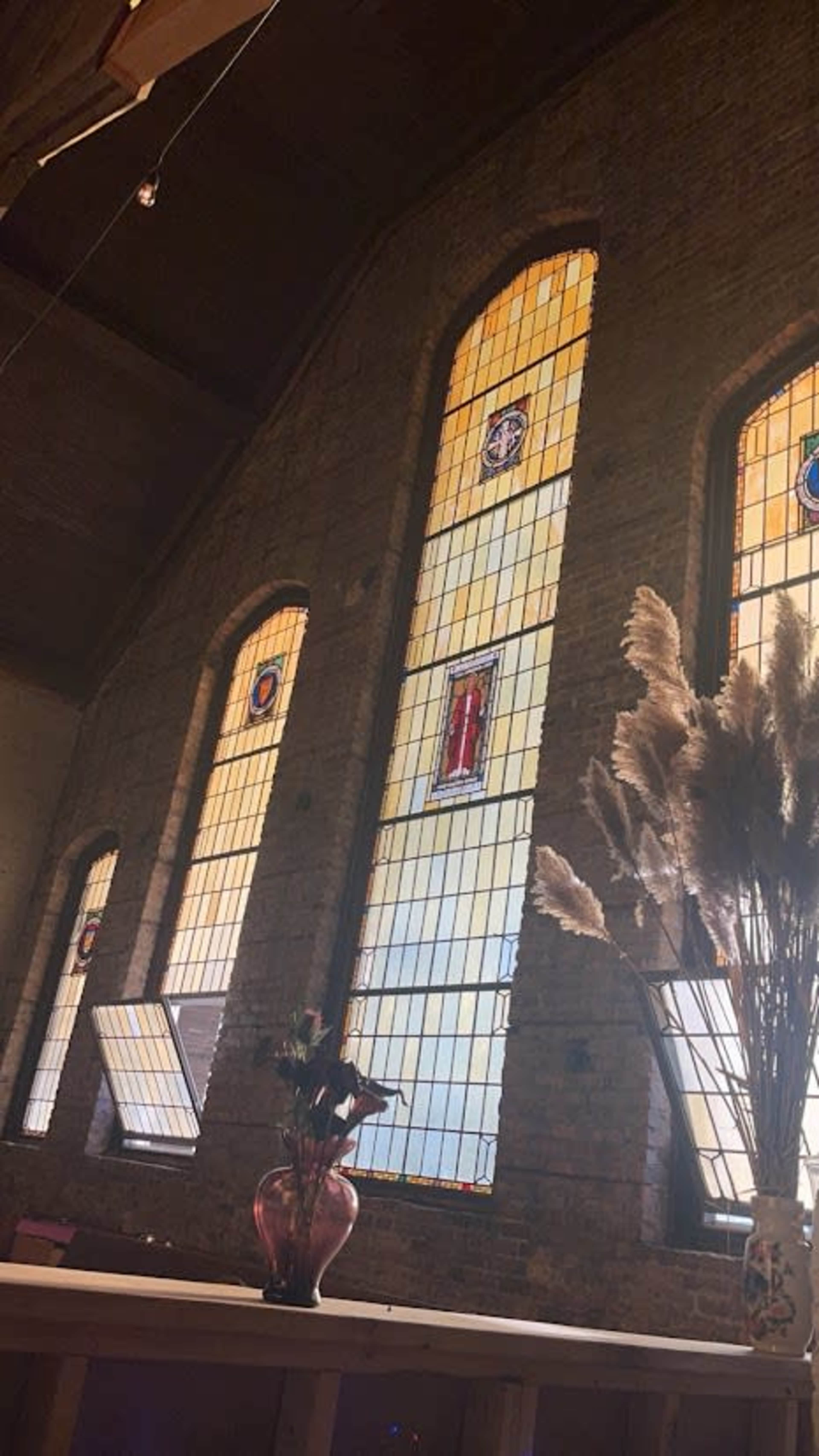 The image shows a room with three tall stained glass windows and a decorative arrangement of dried plants in a vase on a table in the foreground.