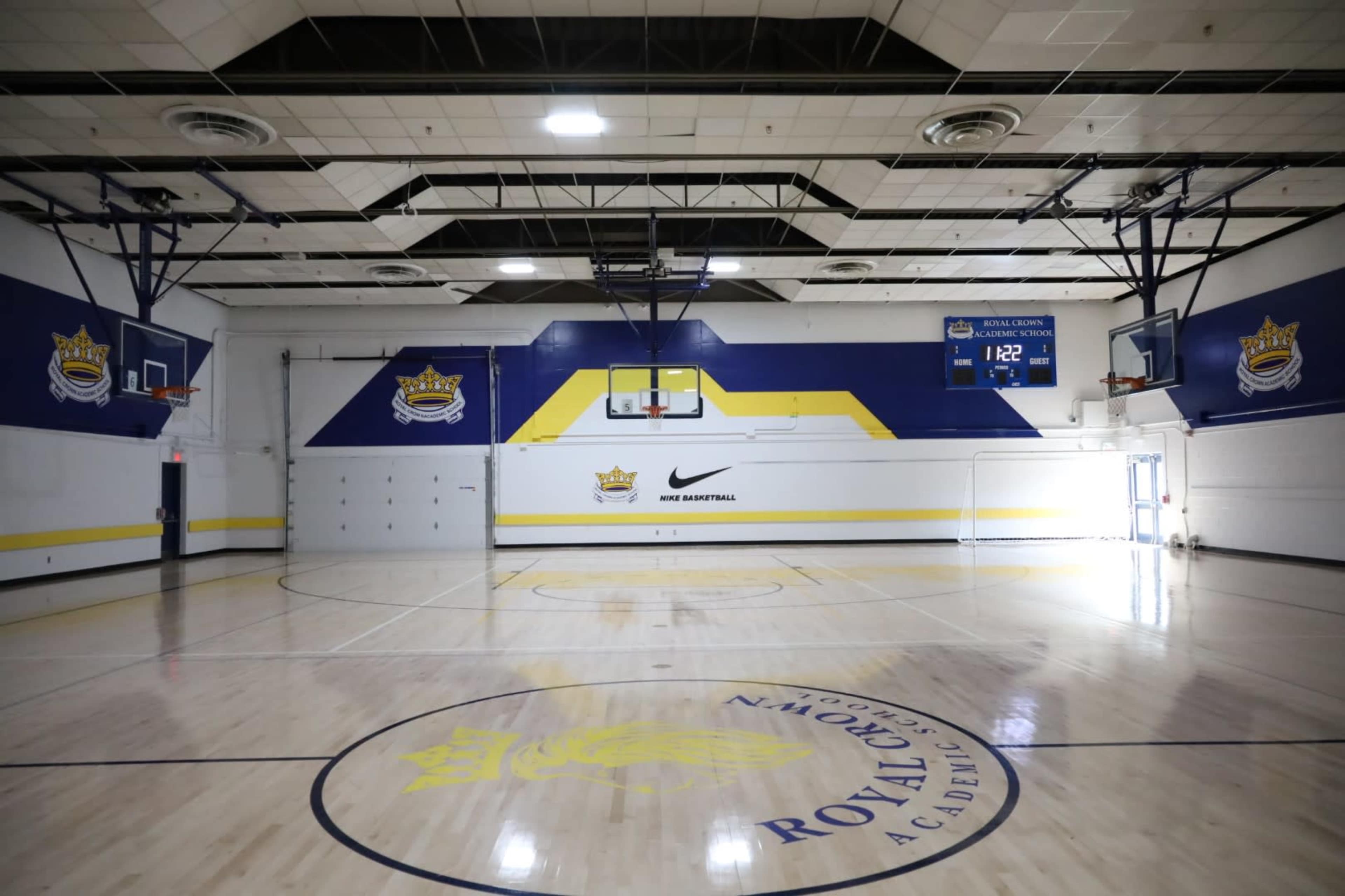 The image shows a basketball gymnasium with hardwood floors, multiple basketball hoops, and a scoreboard displaying the time.