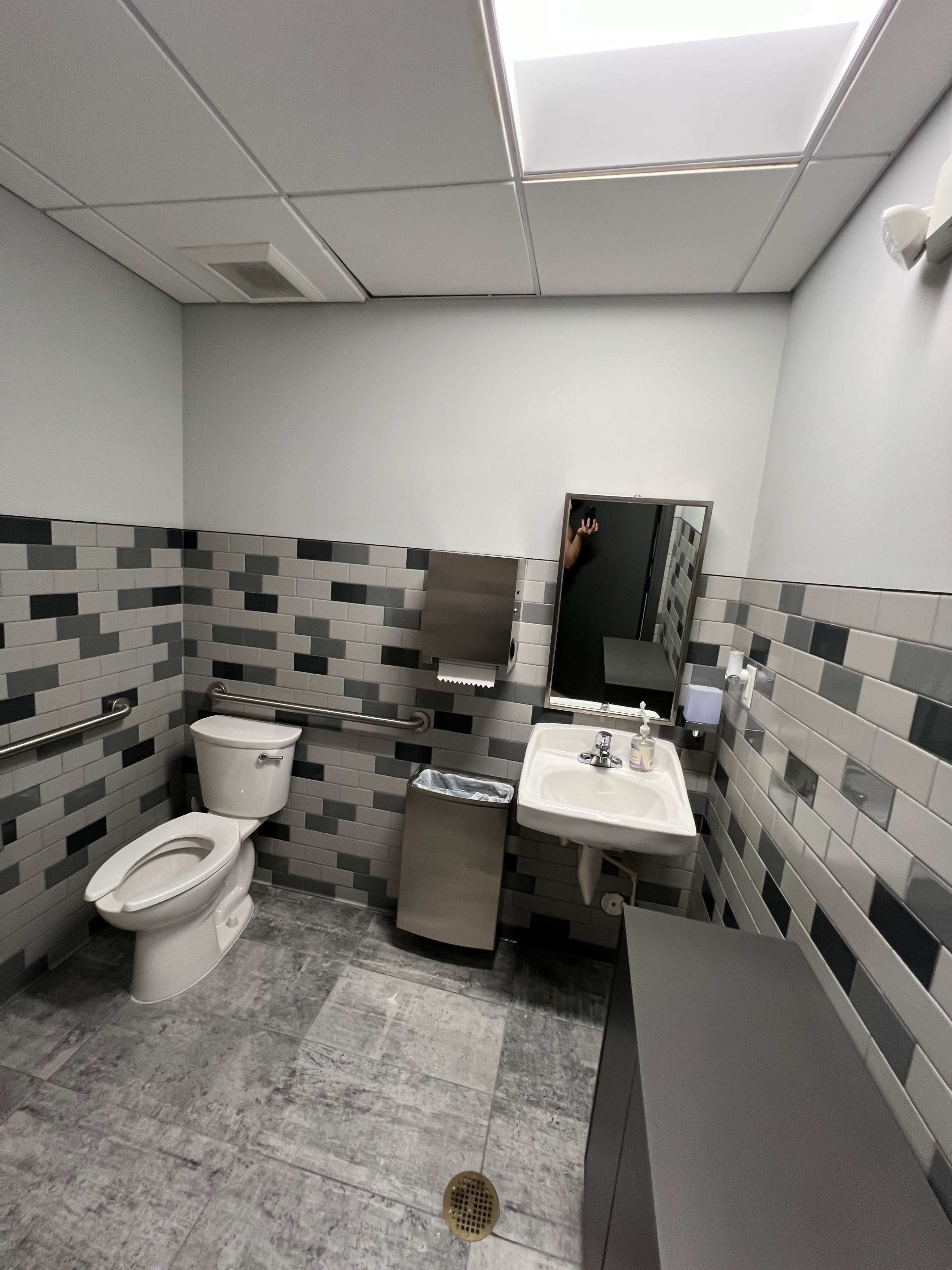 A modern bathroom with a toilet, a sink, a mirror, a trash can, and tiled walls in gray tones.