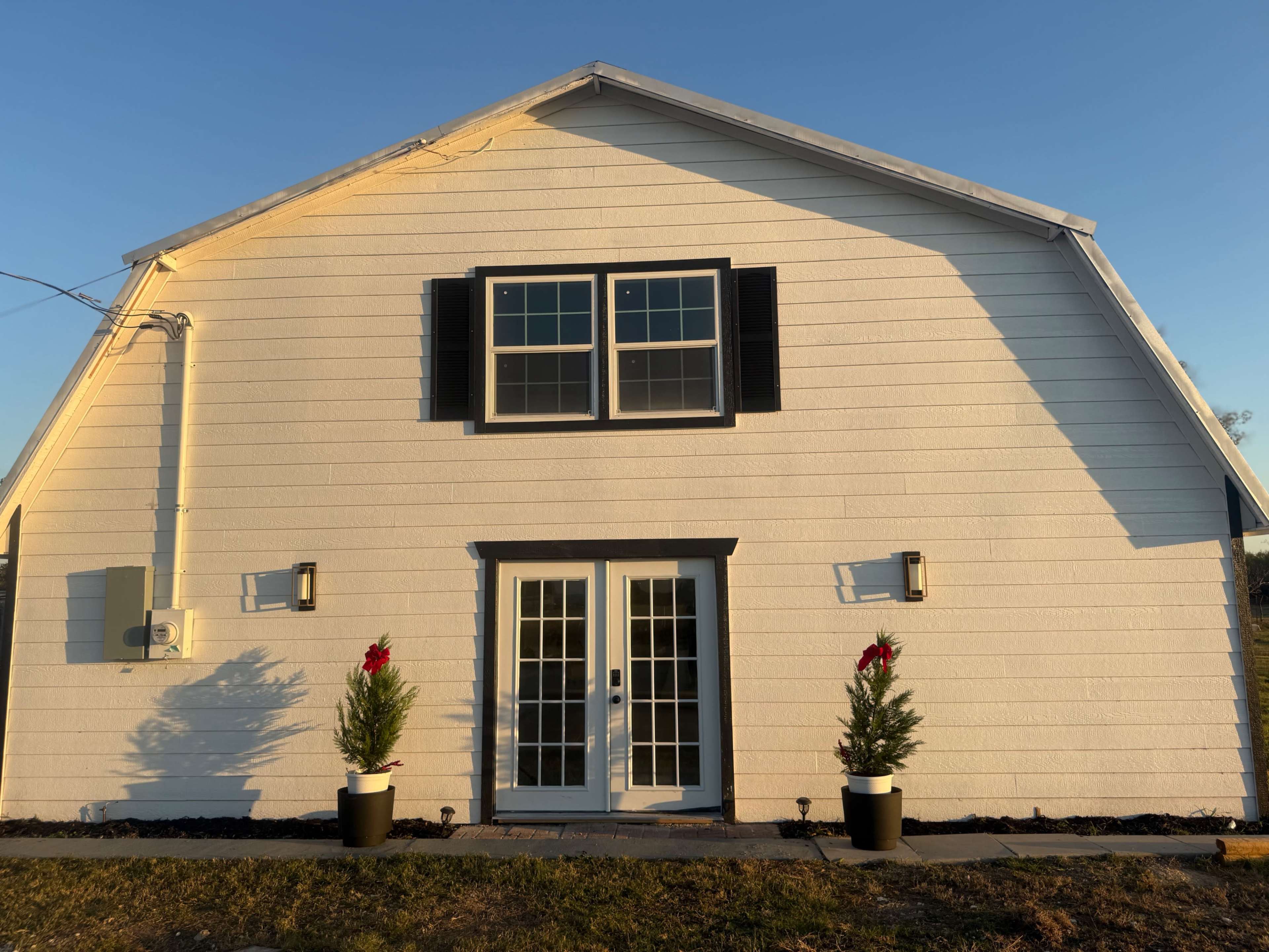 The image shows a white, gable-roofed house with black shutters, featuring a set of double doors flanked by potted greenery adorned with red accents.