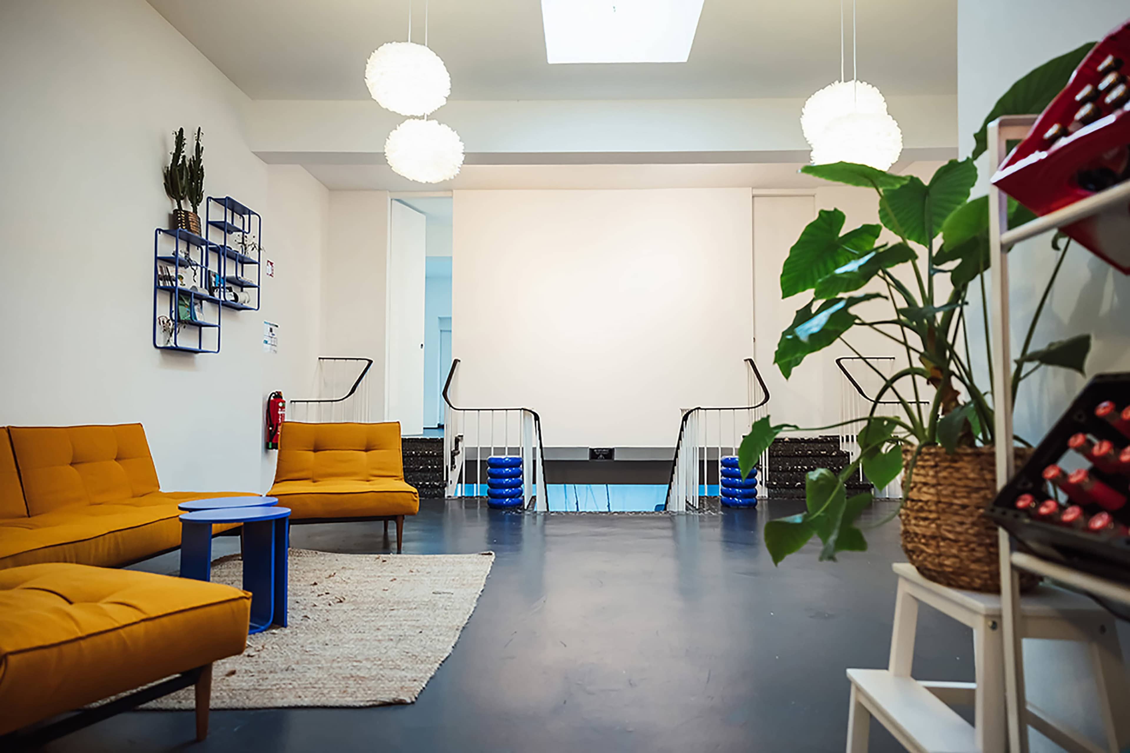 The image shows a modern room with two orange sofas, a round blue table, and tall potted plants, with a pool area visible in the background.