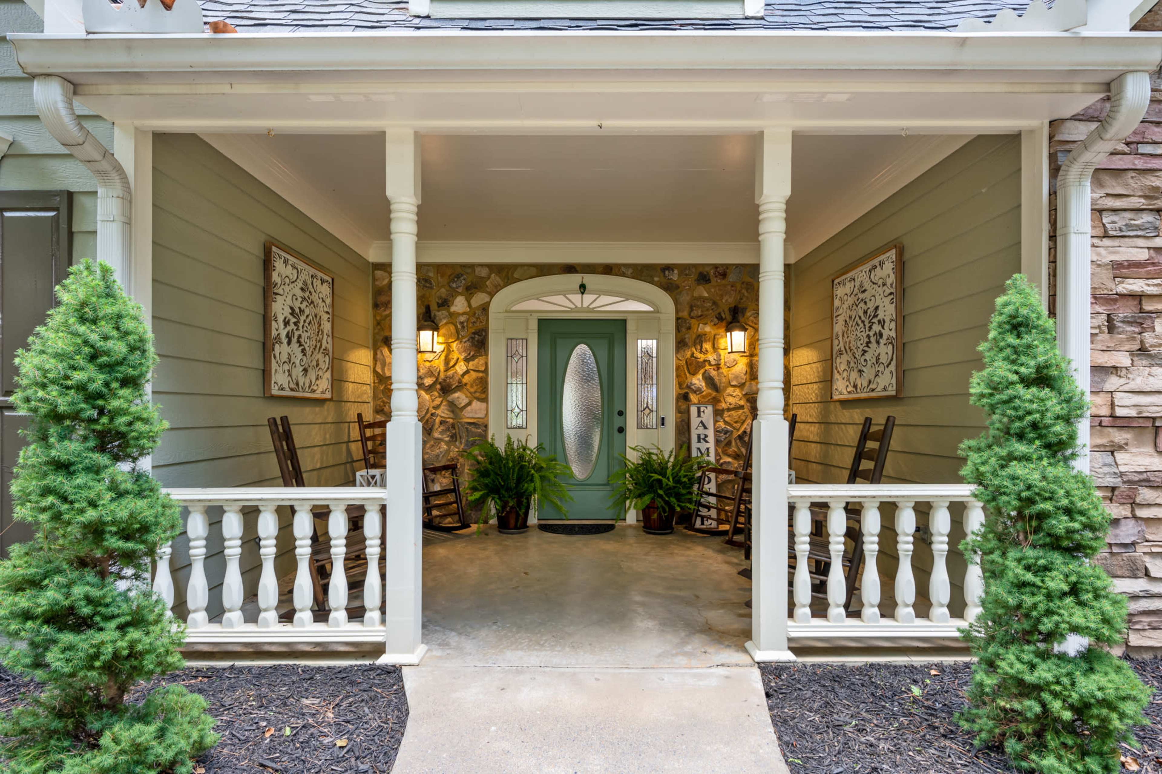 A covered porch with rocking chairs, decorative wall art, and potted plants, framed by two small evergreen shrubs.