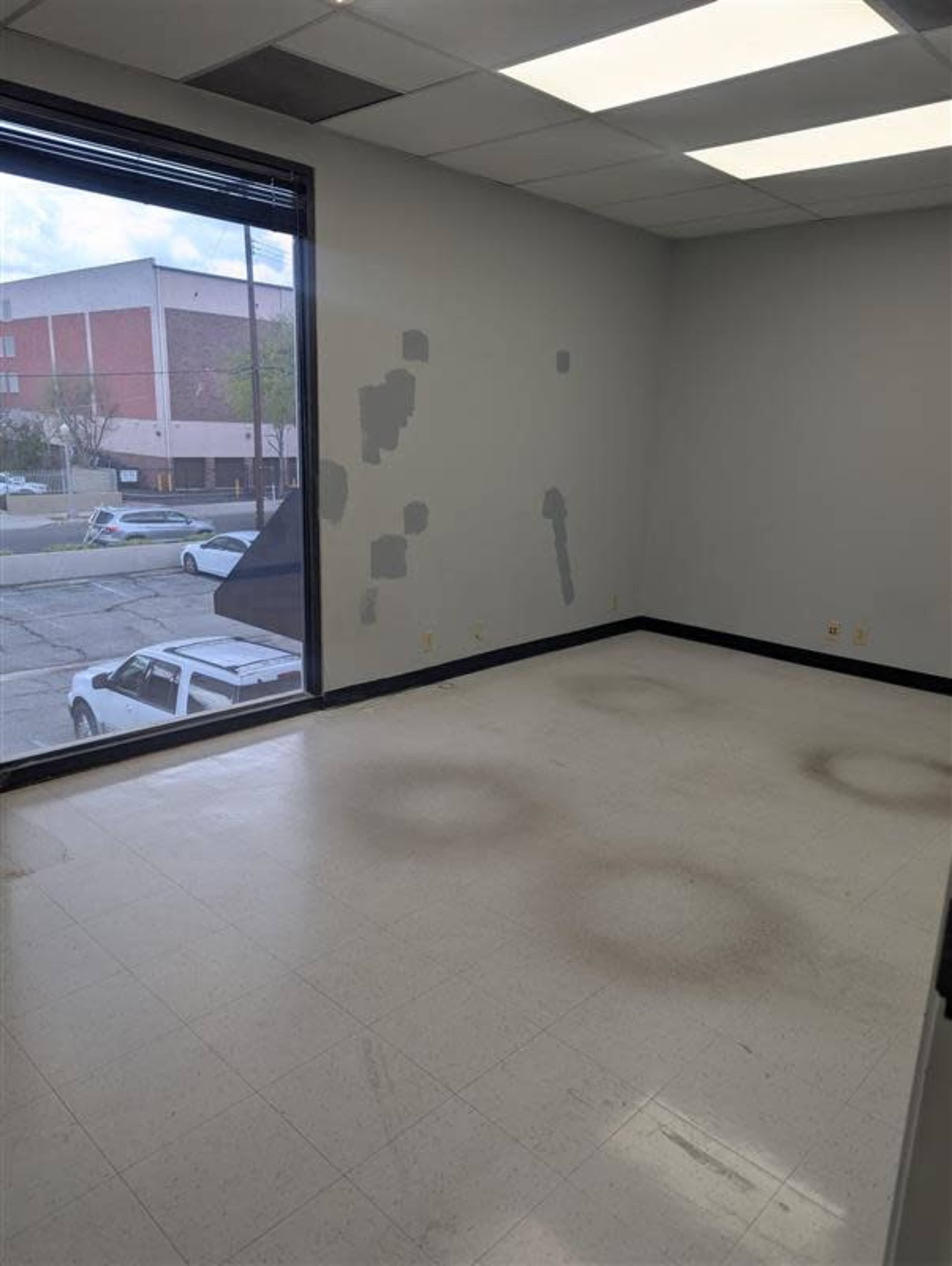The image shows a vacant office room with a large window, light-colored walls, and noticeable stains on the floor.
