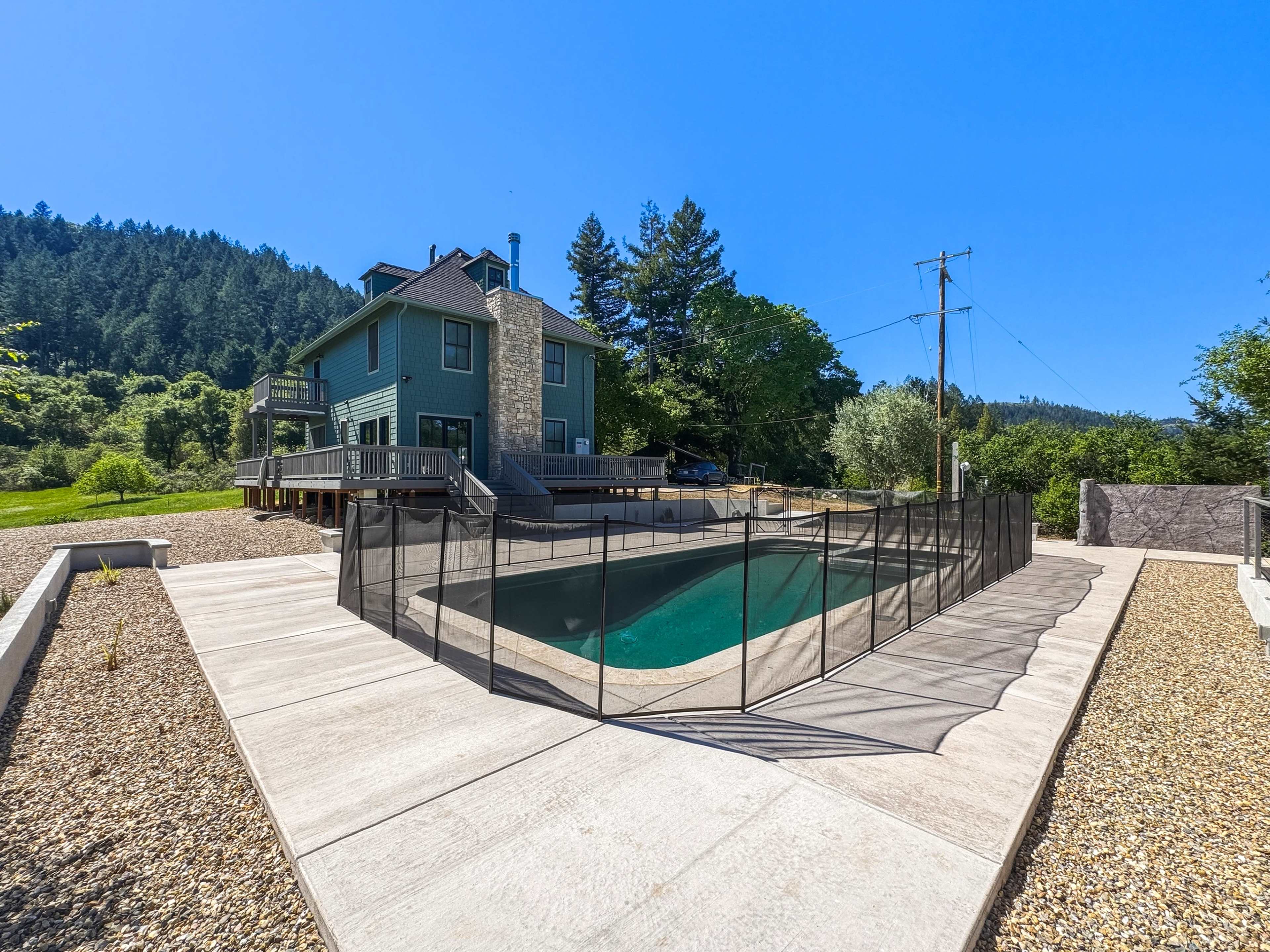 A two-story blue house with a stone chimney overlooks a fenced pool area surrounded by gravel, with a forested hillside in the background.