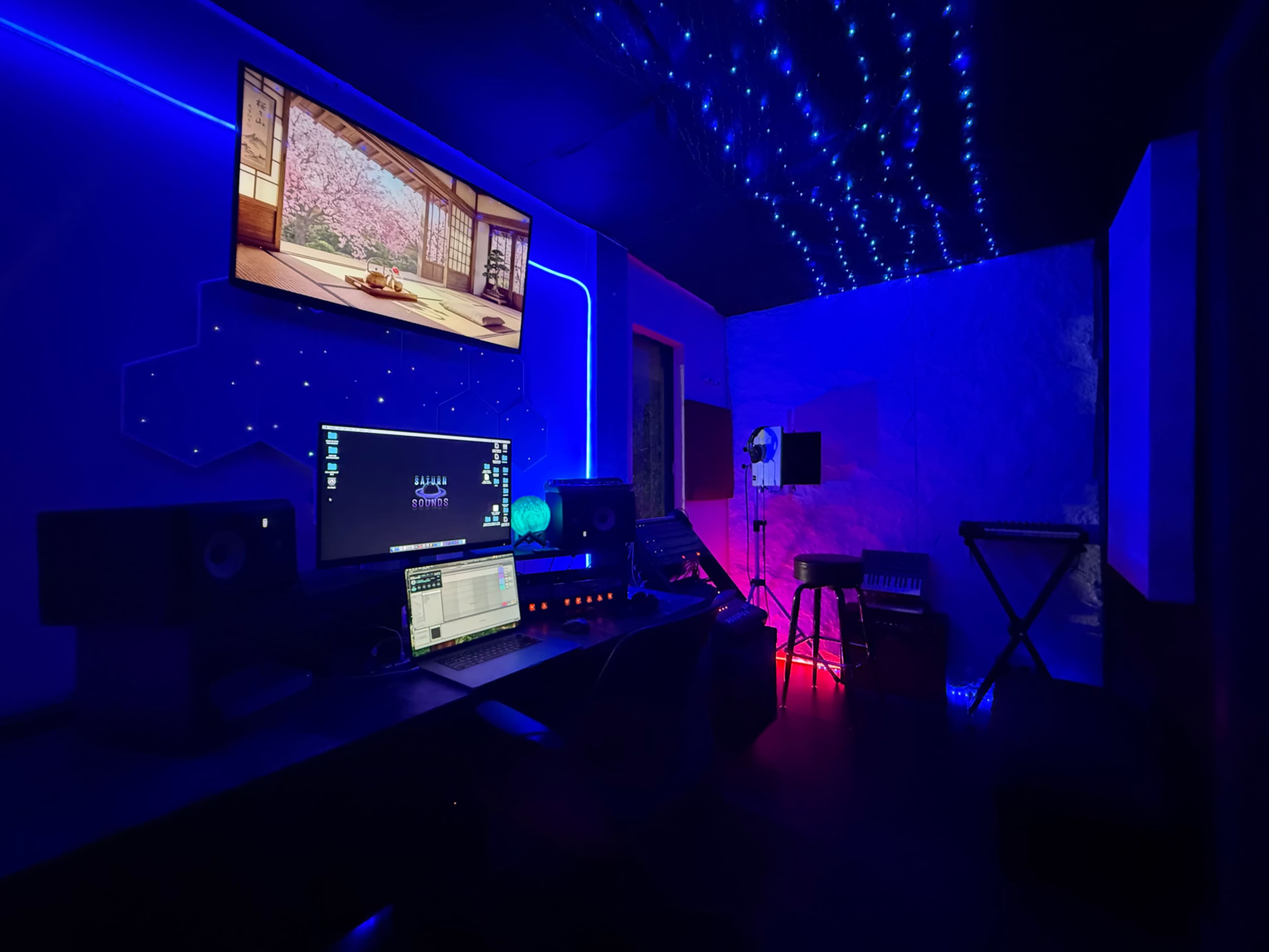 The image shows a modern music studio with a computer setup, speakers, and colorful LED lights illuminating the dark space.