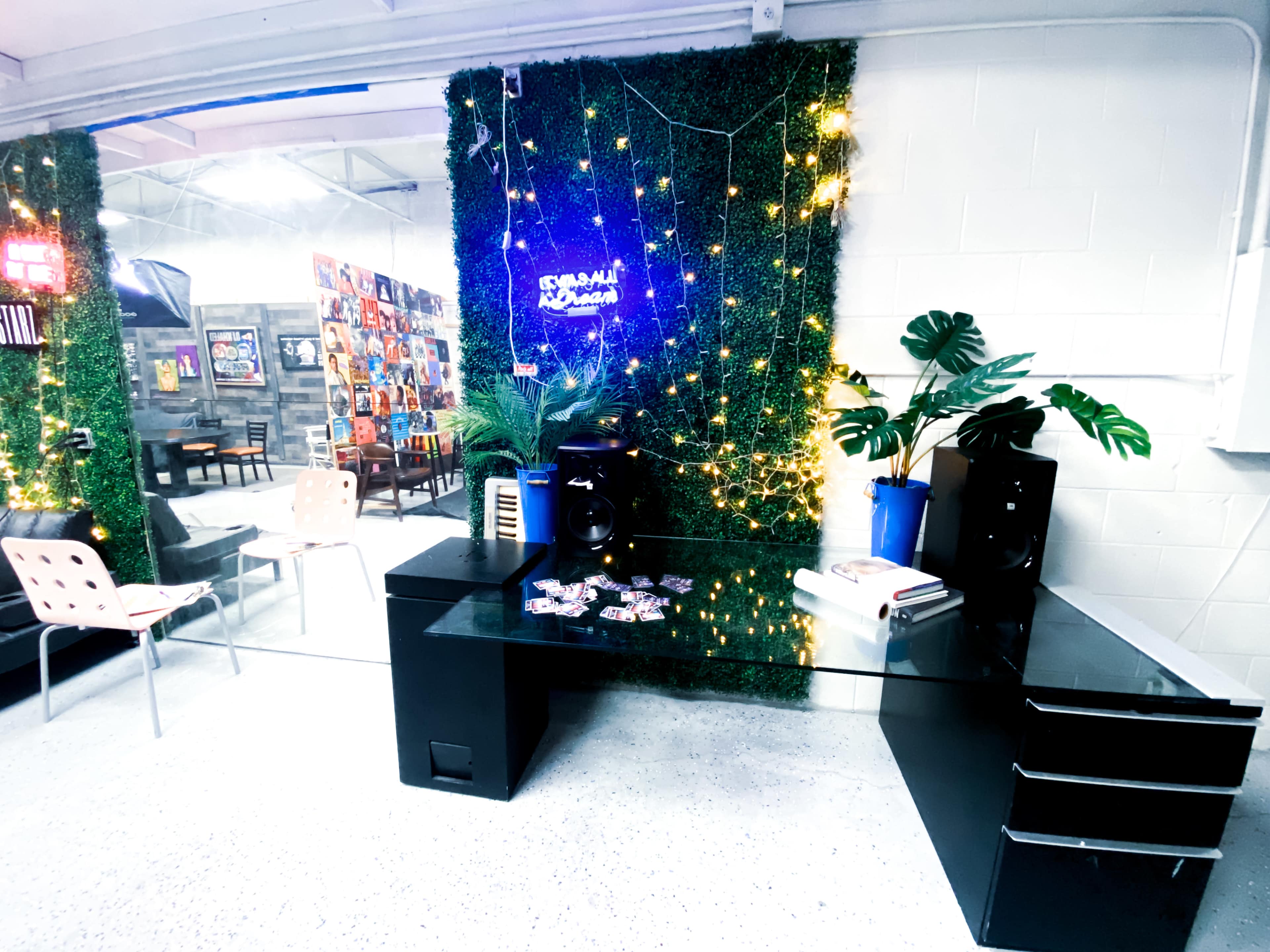 The image shows a modern workspace with a glass desk, an audio speaker, and a wall covered in greenery and string lights.