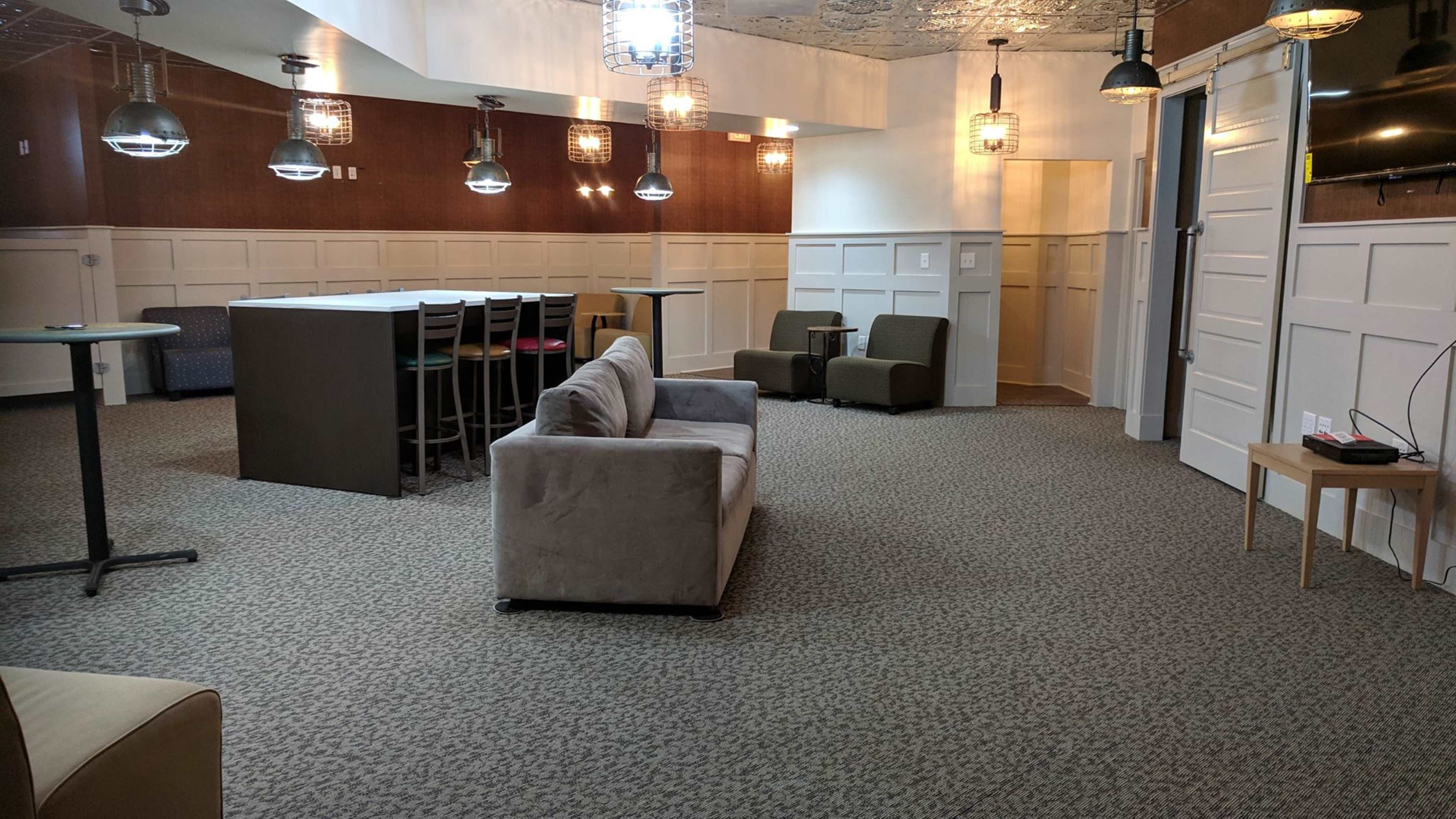 A modern lounge area featuring a gray sofa, two green chairs, a counter with high chairs, and pendant lighting against a paneled wall.