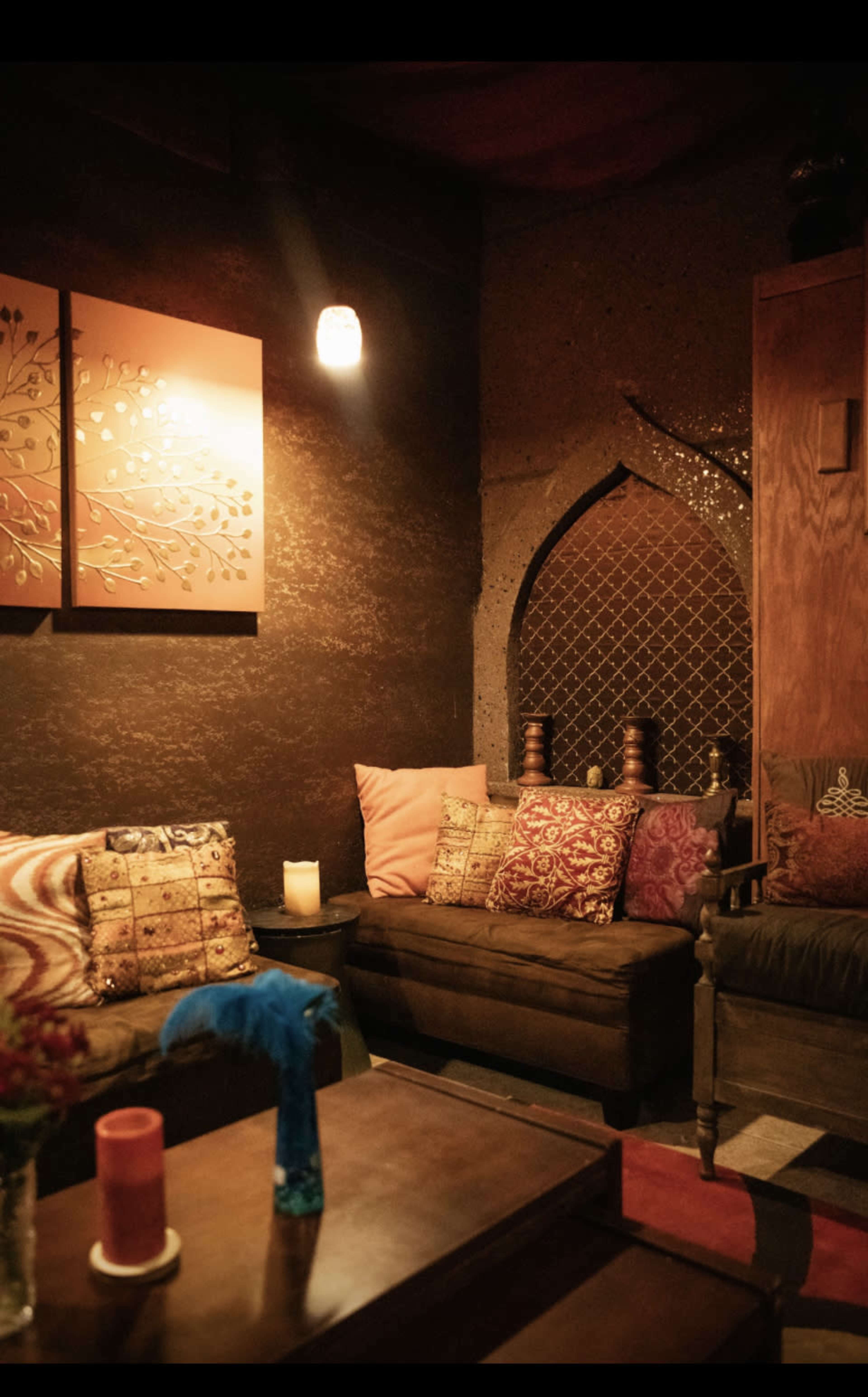 A cozy lounge area with patterned cushions, a wooden table, and soft lighting, featuring an arched window in the background.