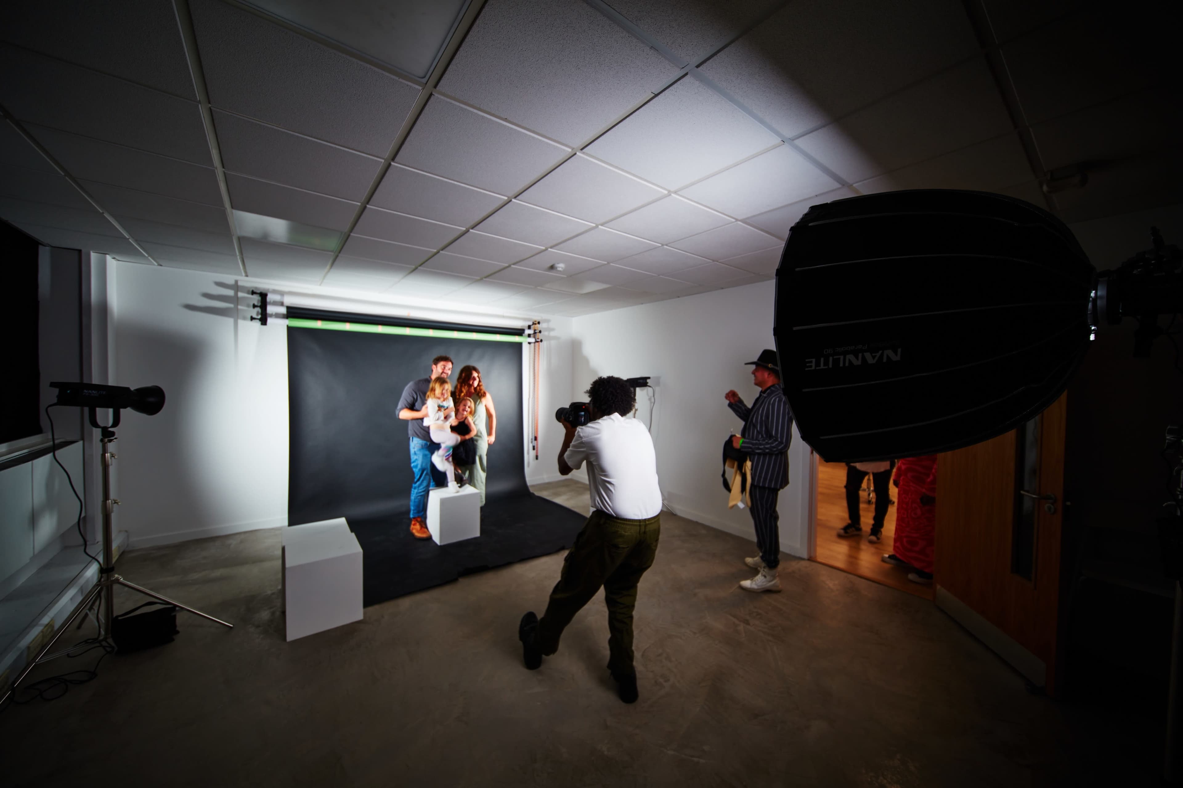 A photographer captures a couple posing on white blocks in a photo studio with a black backdrop and softbox lighting.