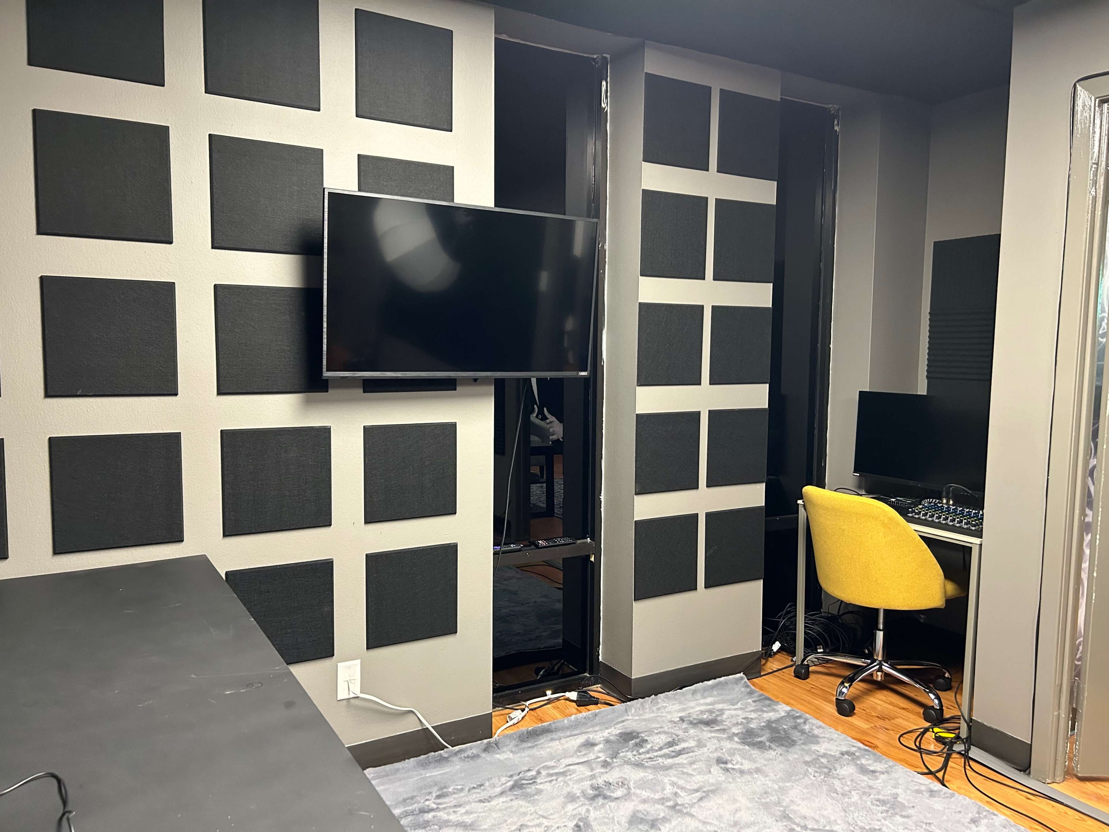 A recording studio with soundproofed walls, a television mounted on one wall, and a desk with a computer and a yellow chair in the corner.