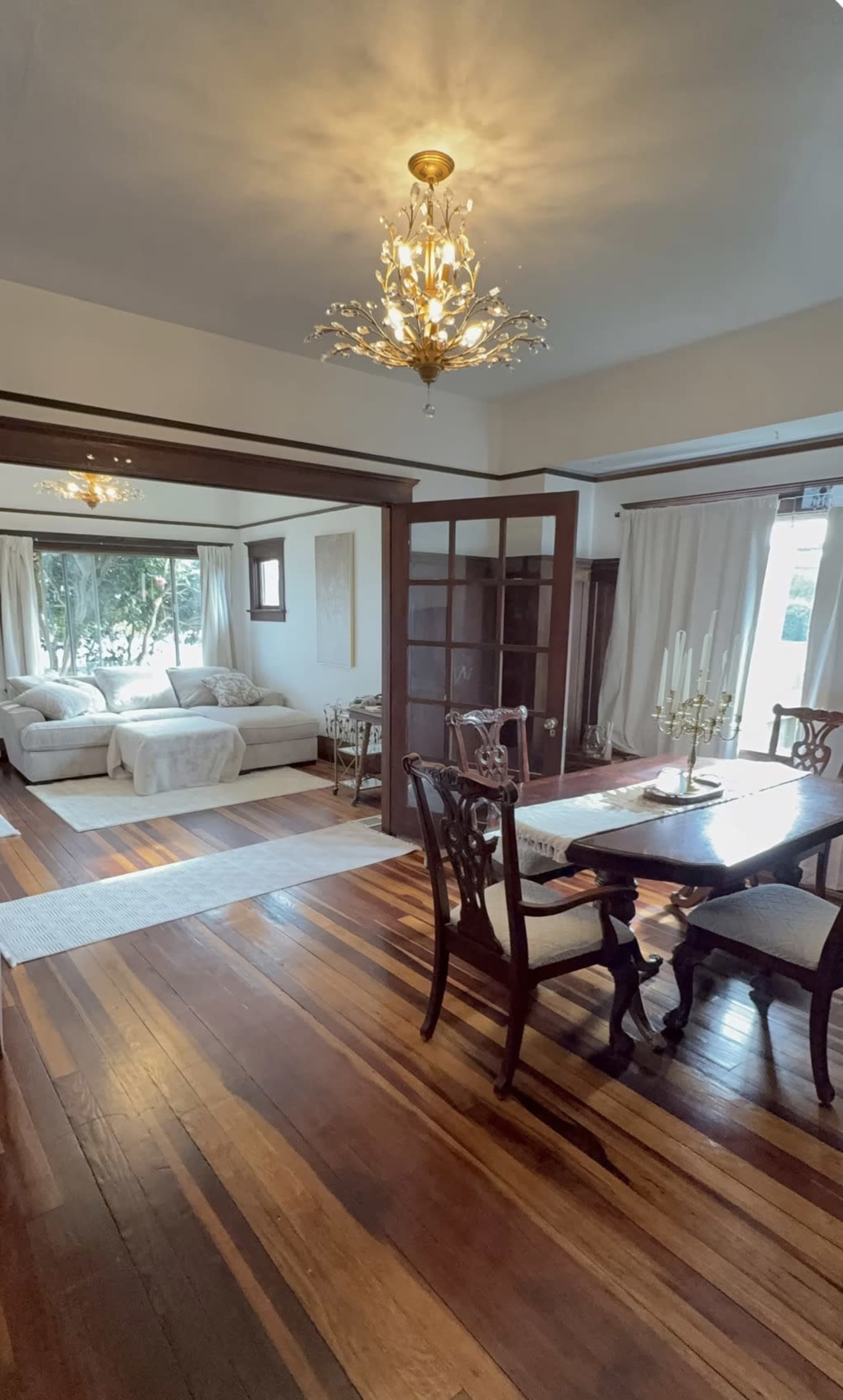 The image shows a dining area with a wooden table and chairs, leading into a living room featuring a light-colored sofa and large windows draped with sheer curtains.