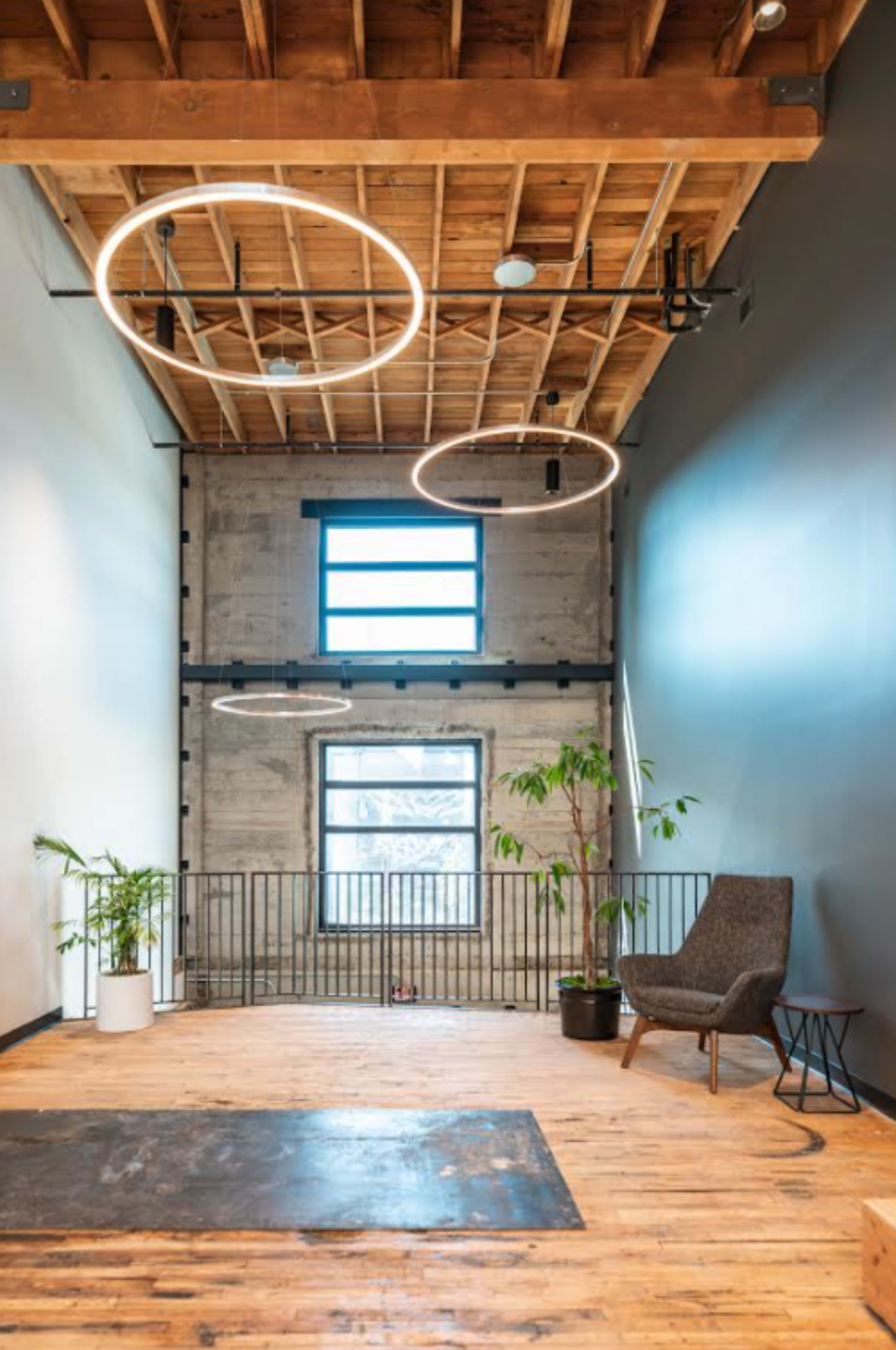 A spacious interior of an office with high ceilings, wooden beams, circular lights, large windows, and a green plant next to a gray chair.