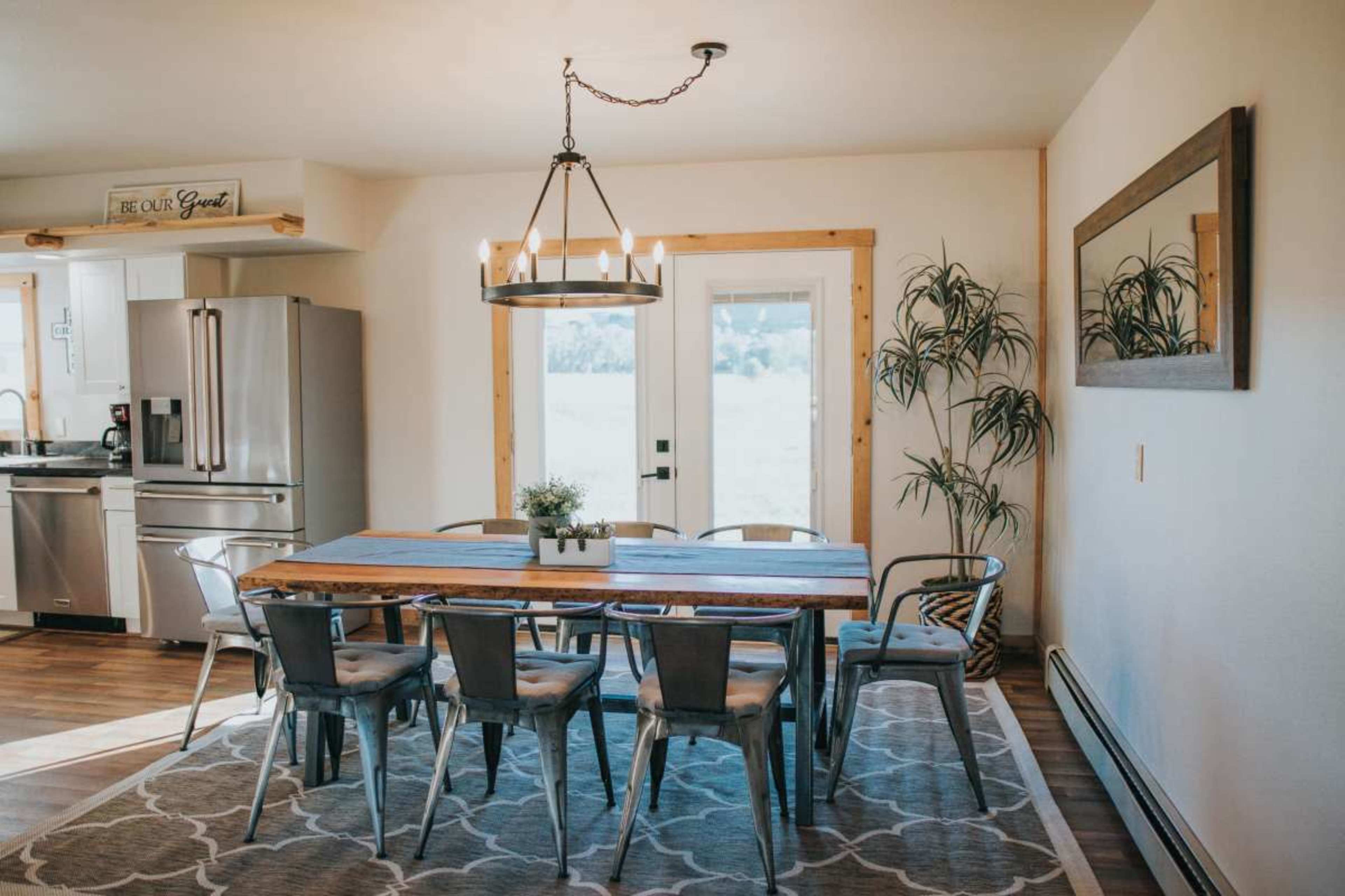 A spacious dining area features a large wooden table surrounded by metal-framed chairs, with a chandelier overhead and sliding glass doors leading to an outdoor view.