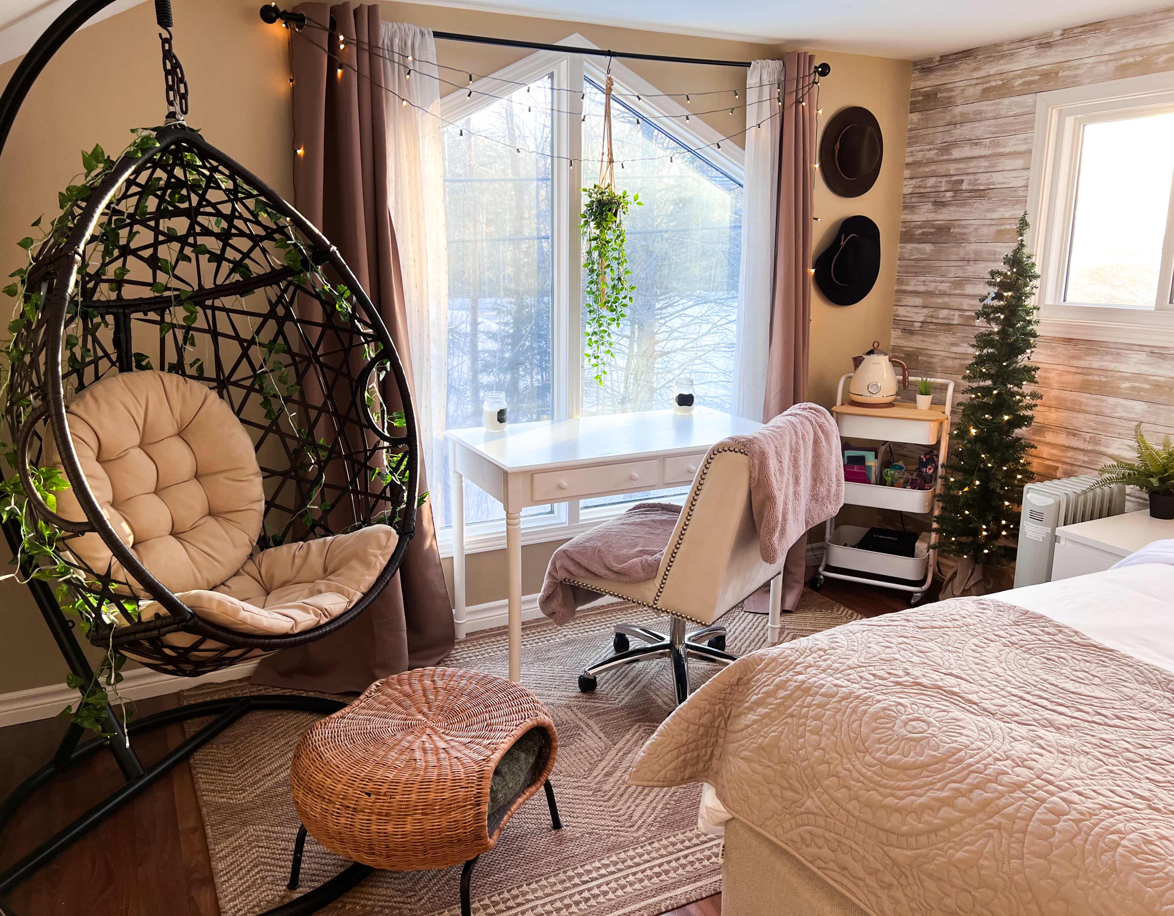 A cozy room featuring a hanging chair, a desk with a chair, and a small tree, all arranged near a large window with natural light.
