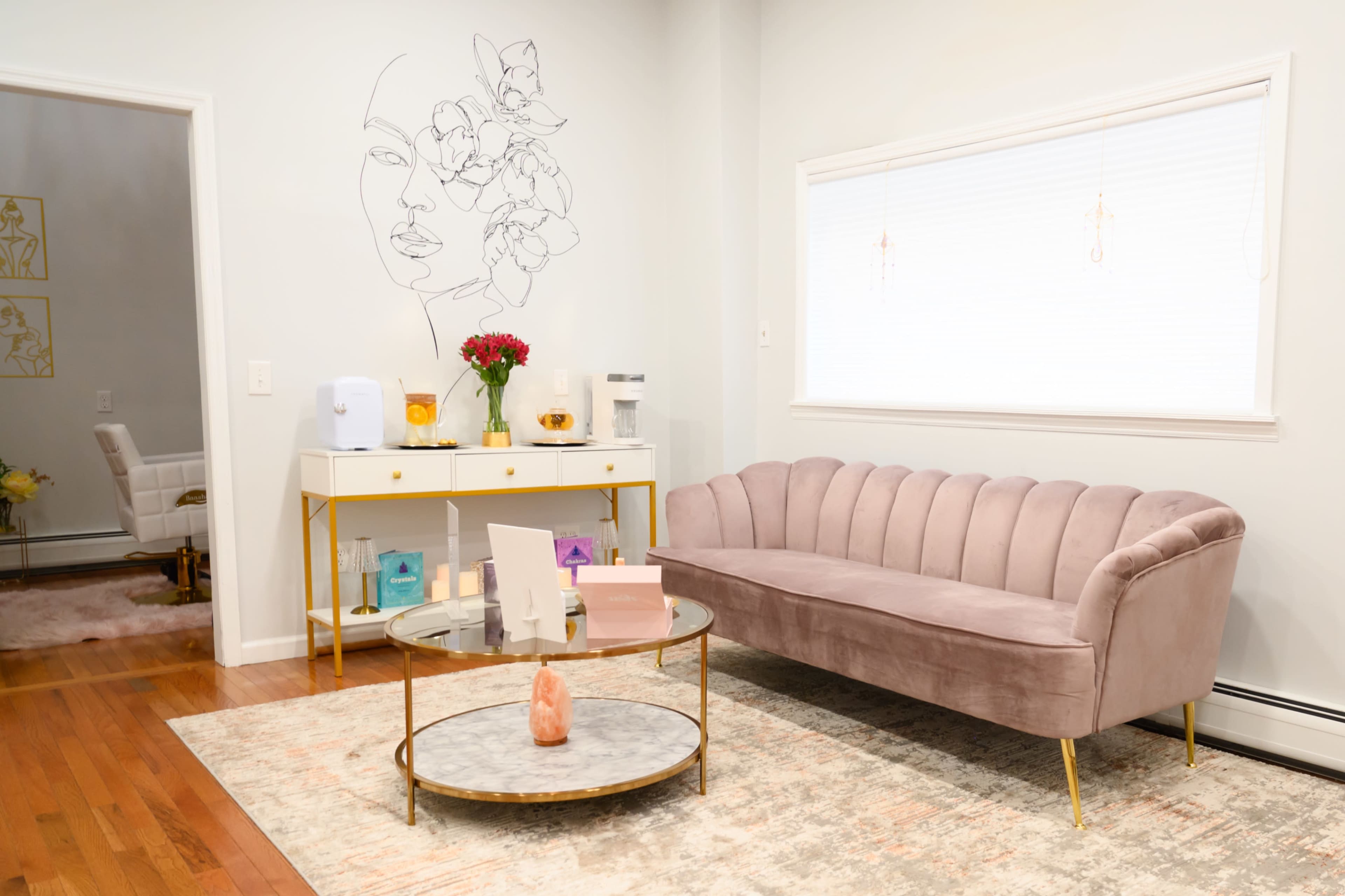 The image shows a modern, softly-lit room featuring a plush pink sofa, a round marble coffee table, and a decorative wall art piece of a face with flowers.