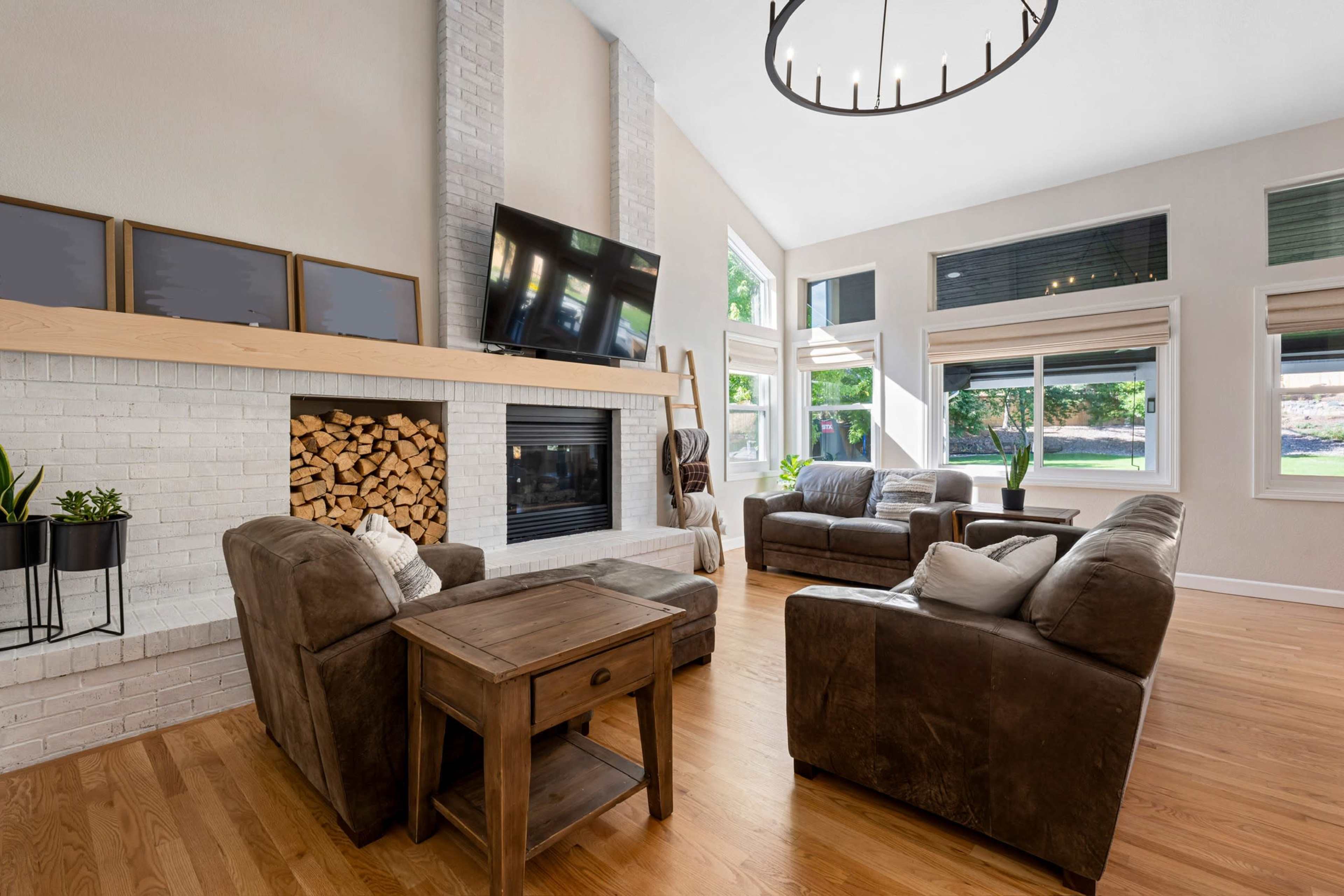 The image depicts a modern living room with two sofas, a coffee table, a fireplace adorned with stacked logs, and large windows allowing natural light to enter.