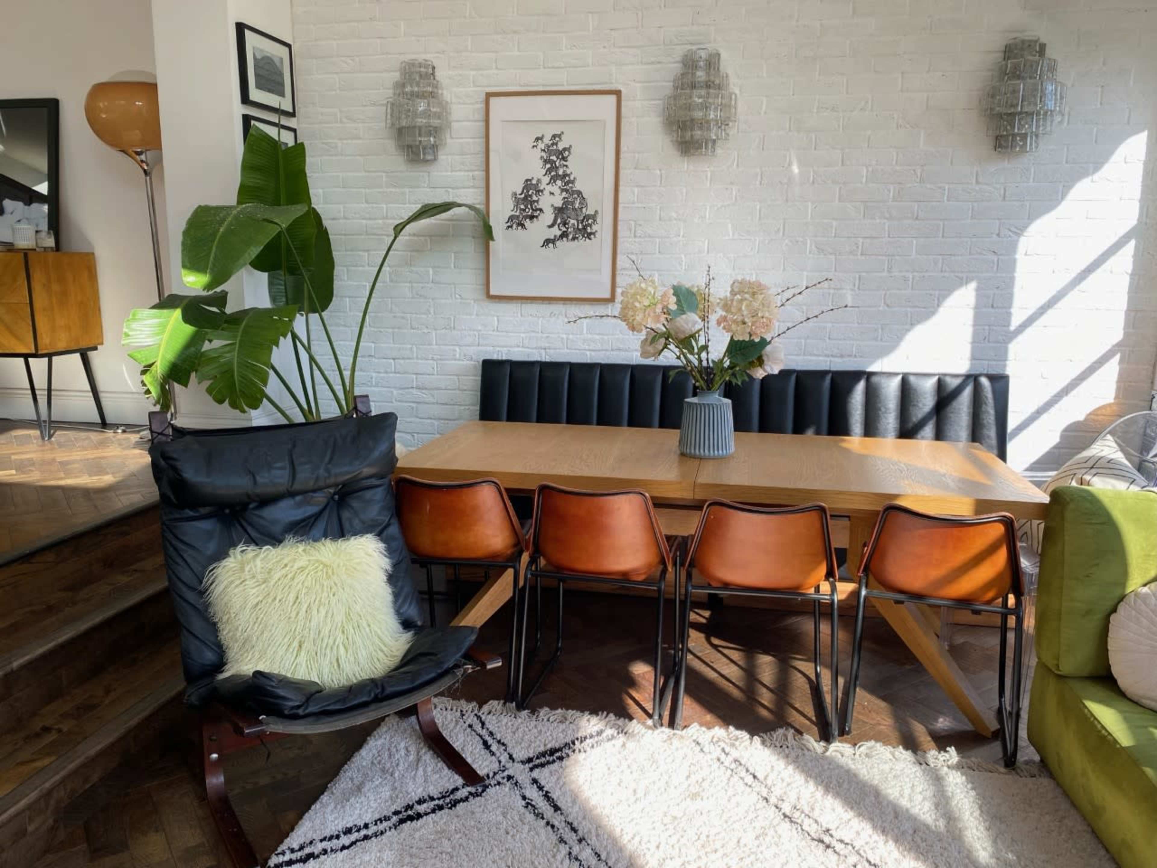 The image shows a modern dining area with a wooden table surrounded by brown chairs, a green couch, and a potted plant in a black chair, illuminated by natural light.