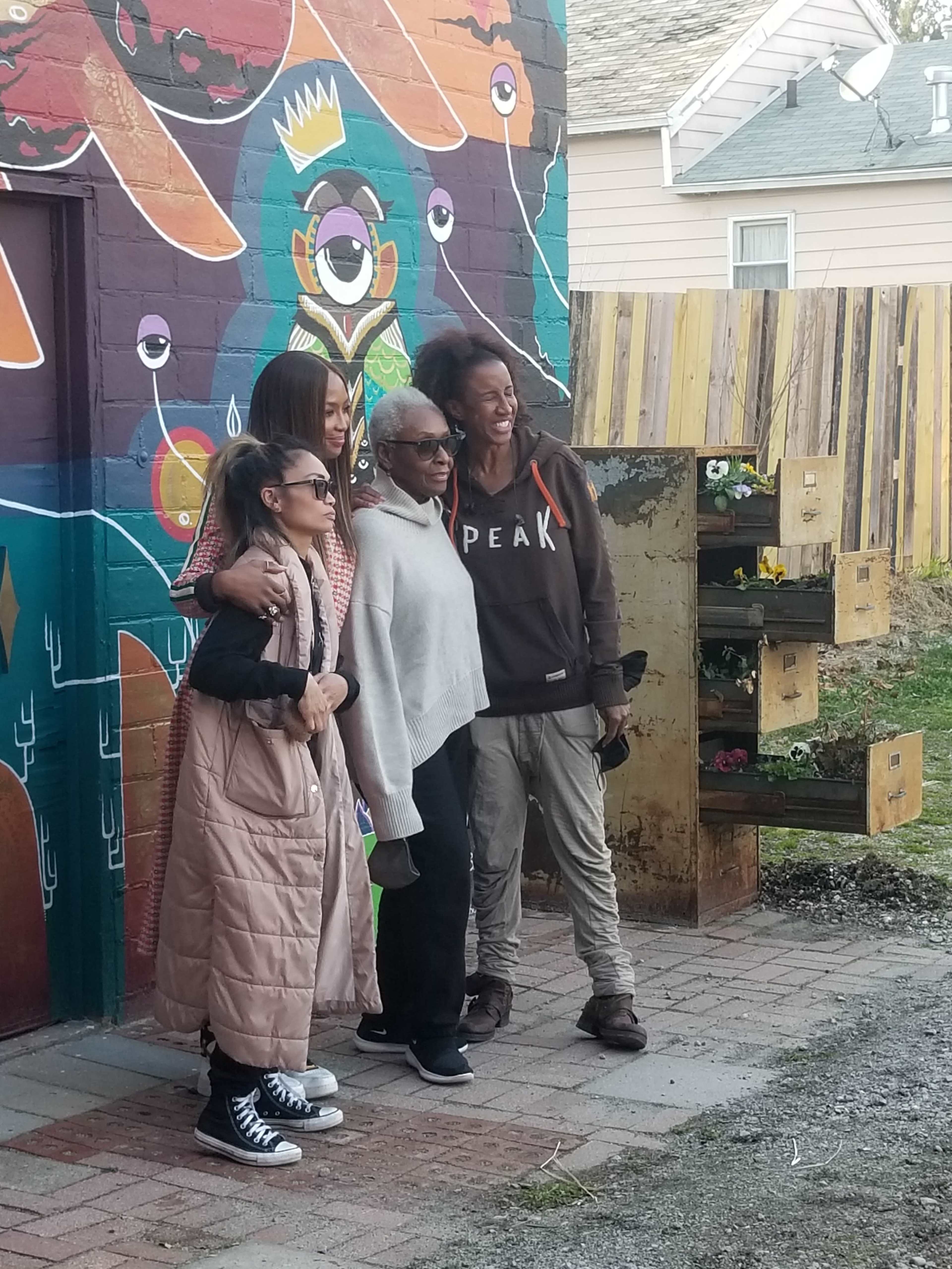 Four individuals pose for a photo in front of a vibrant mural while standing in an outdoor area with wooden fencing and planters.