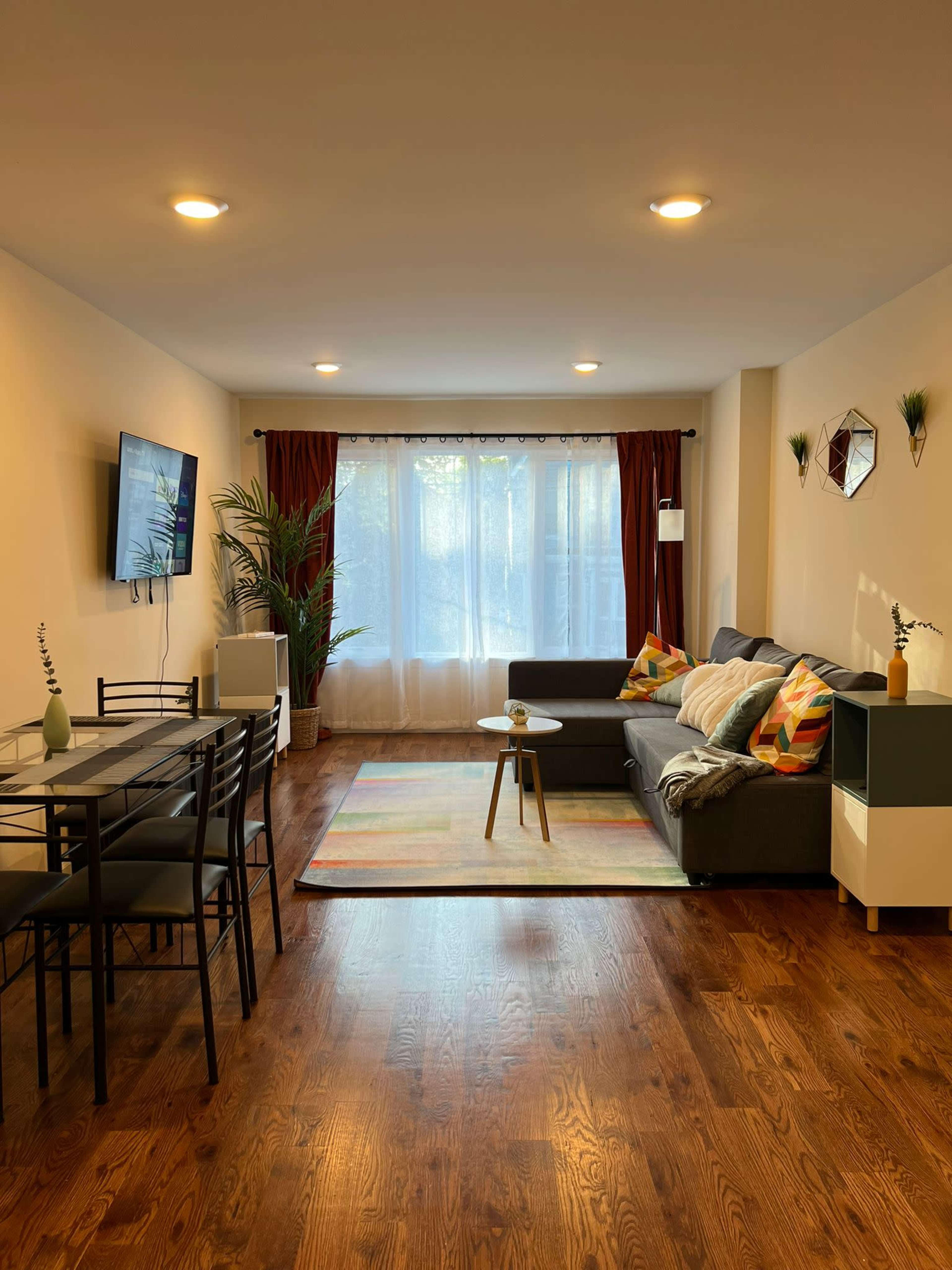 A modern living room features a gray sofa, a dining table with chairs, and large windows draped with curtains, allowing natural light to fill the space.