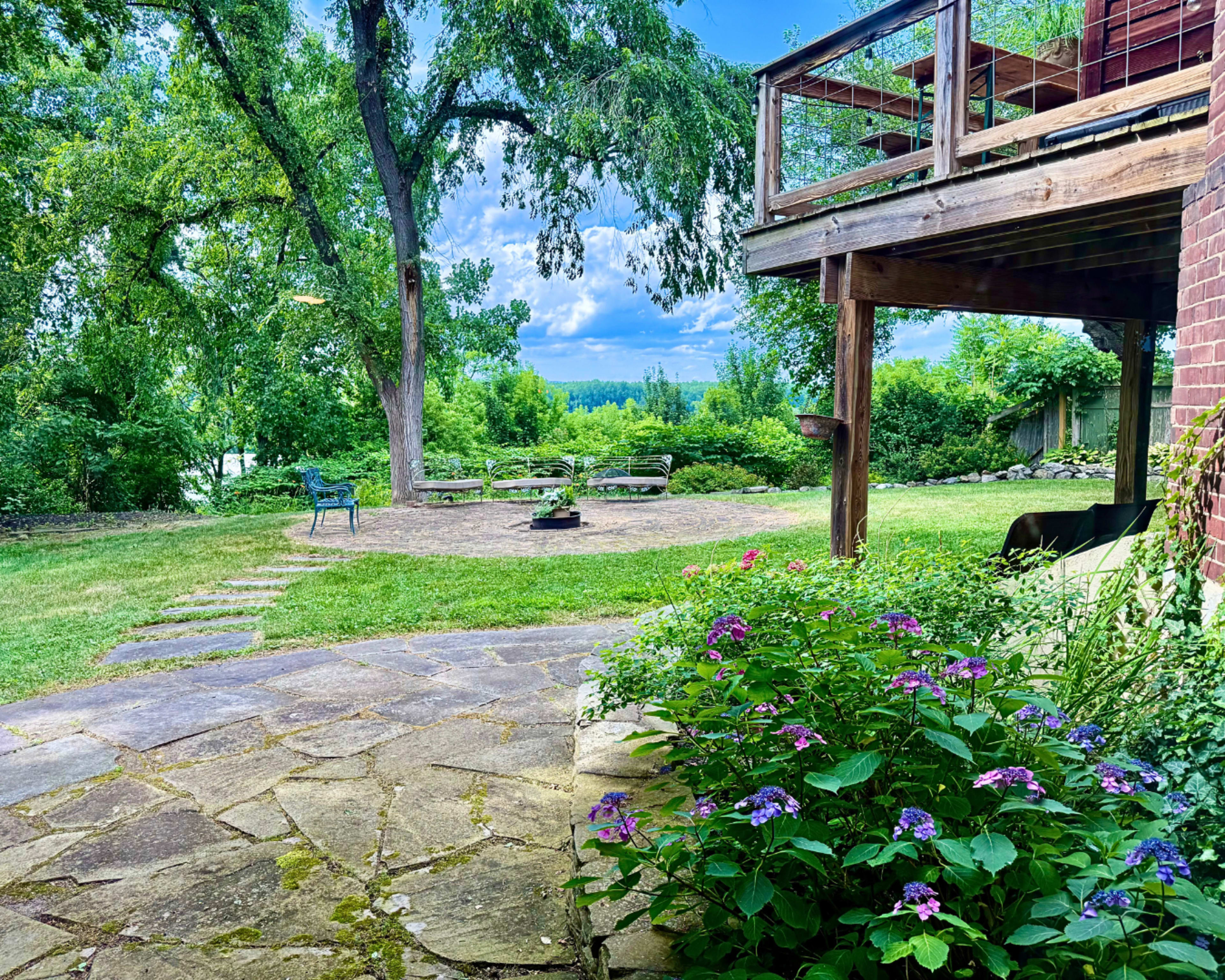 The image shows a stone path leading through a grassy area with flowers to a patio beneath a wooden deck, surrounded by trees and a view of the landscape beyond.