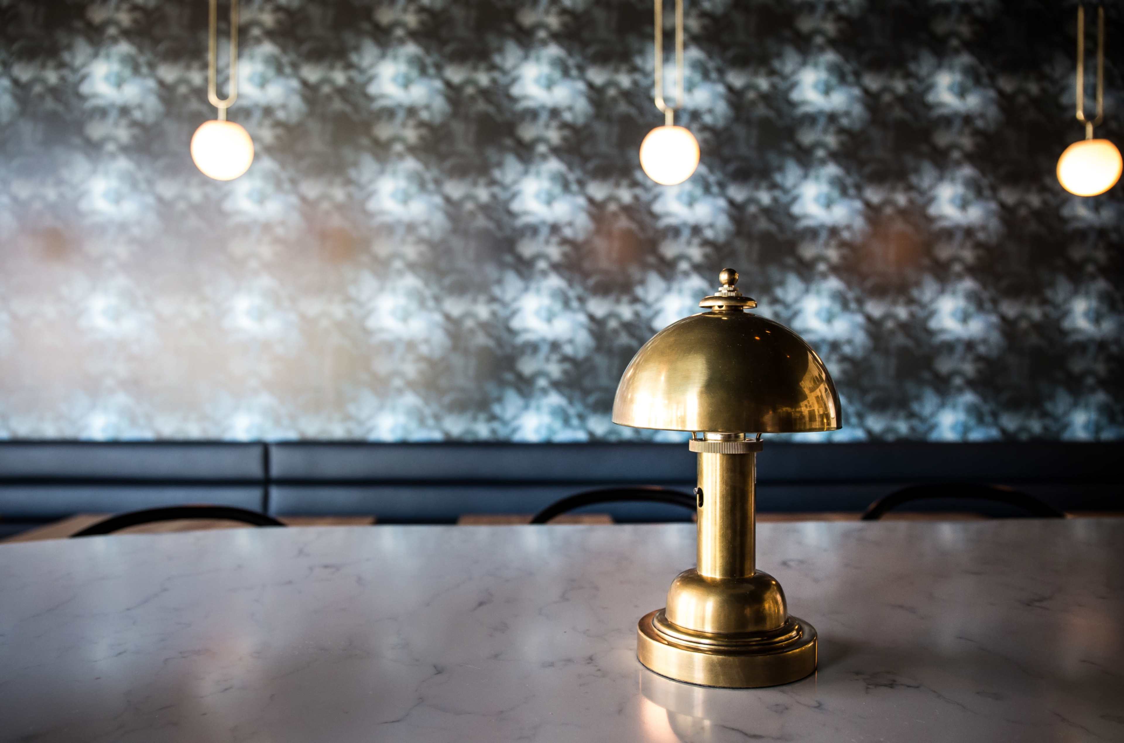 A brass lamp sits on a marble table in a setting featuring a patterned wallpaper and pendant lights in the background.