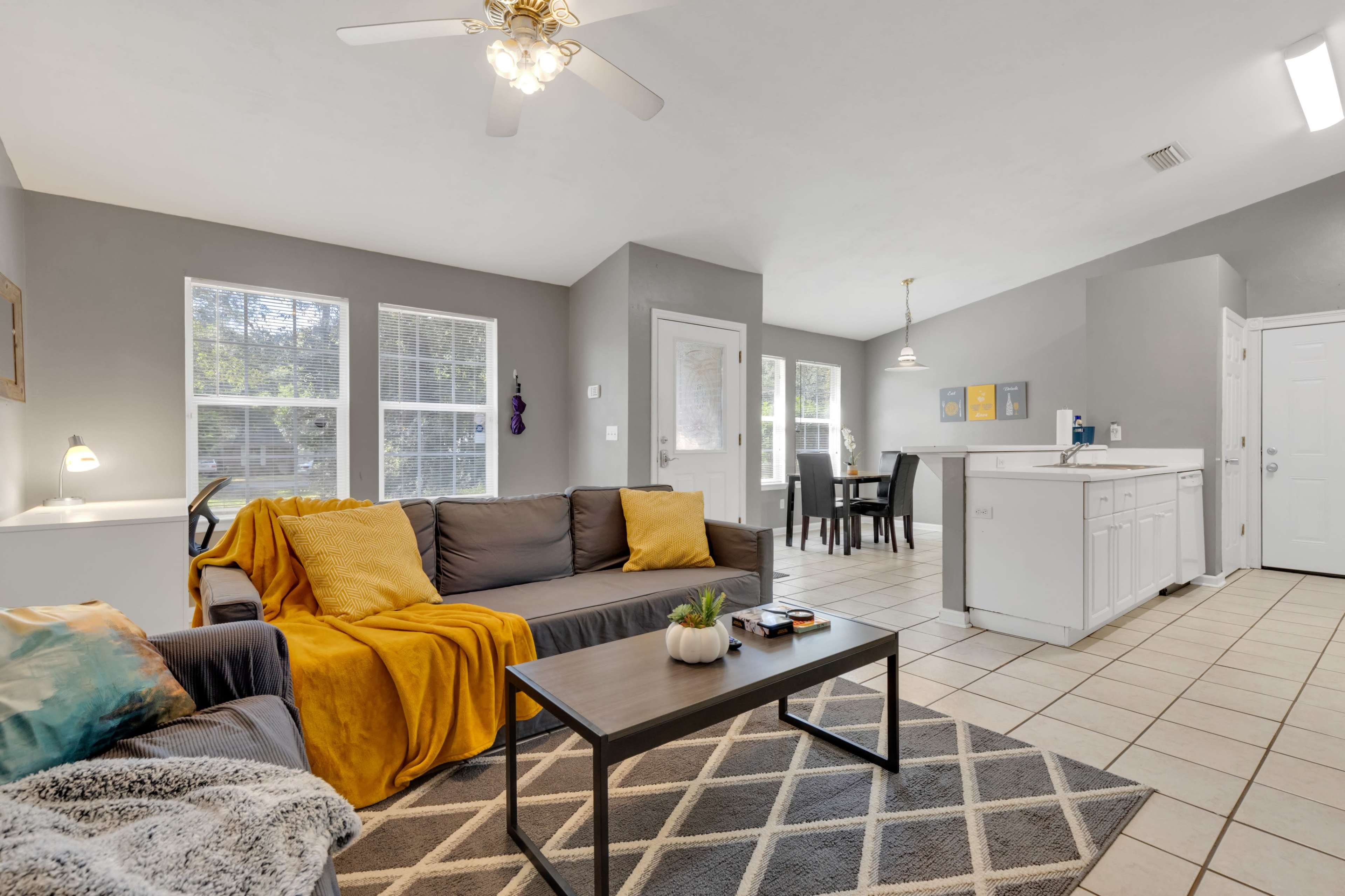 A living room with a grey couch and yellow throw blanket, a coffee table, and an open kitchen area in a well-lit, modern space.